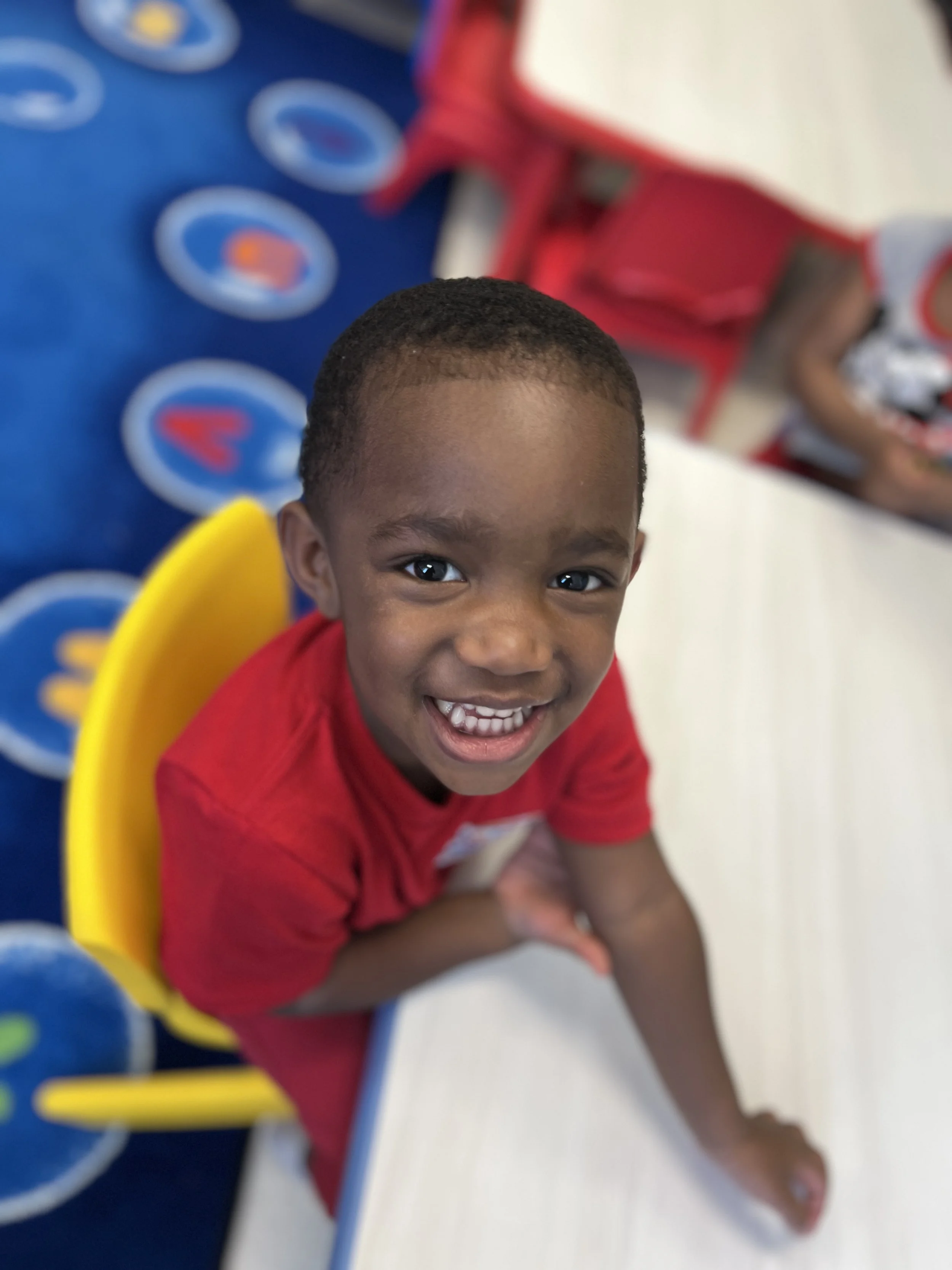Close-up of a young boy with short hair, smiling and looking into the camera. He is wearing a red shirt and sitting on the floor with some children in the background. The background features a blurred colorful play area with various patterns and shap