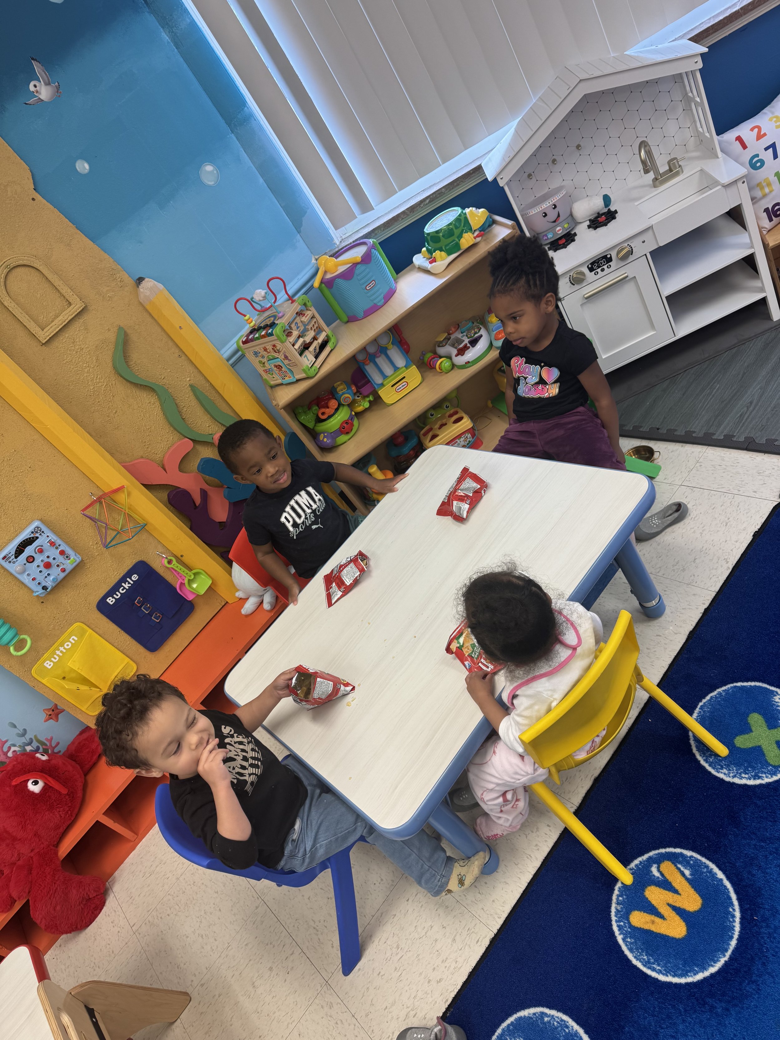Children sitting at a table in a classroom with toys and a play kitchen in the background, engaging in snack time.