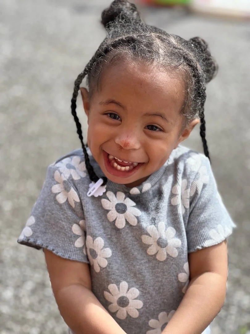 A young girl with dark hair in pigtails, smiling and showing her front teeth, wearing a gray shirt with white daisy patterns, outdoors on a gravel surface.