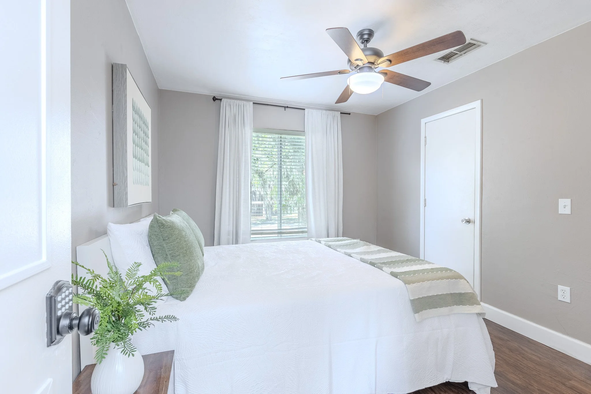 A bedroom with a white bed, white curtains, a window with blinds, and a ceiling fan. The room has neutral-colored walls and wooden flooring.