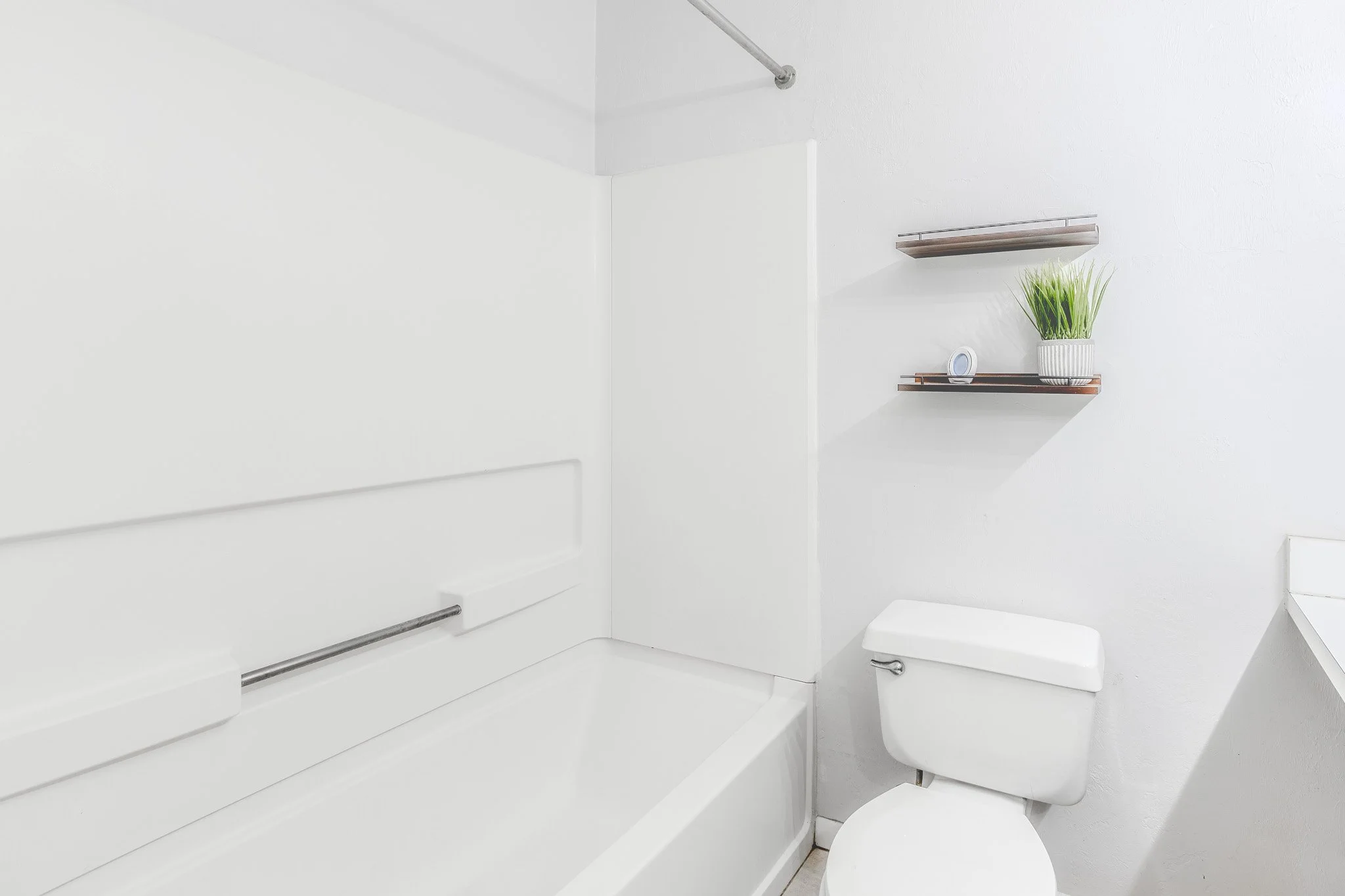A minimalist white bathroom with a toilet, a wall-mounted shelf with a plant, a small round mirror, and a metal towel rack.