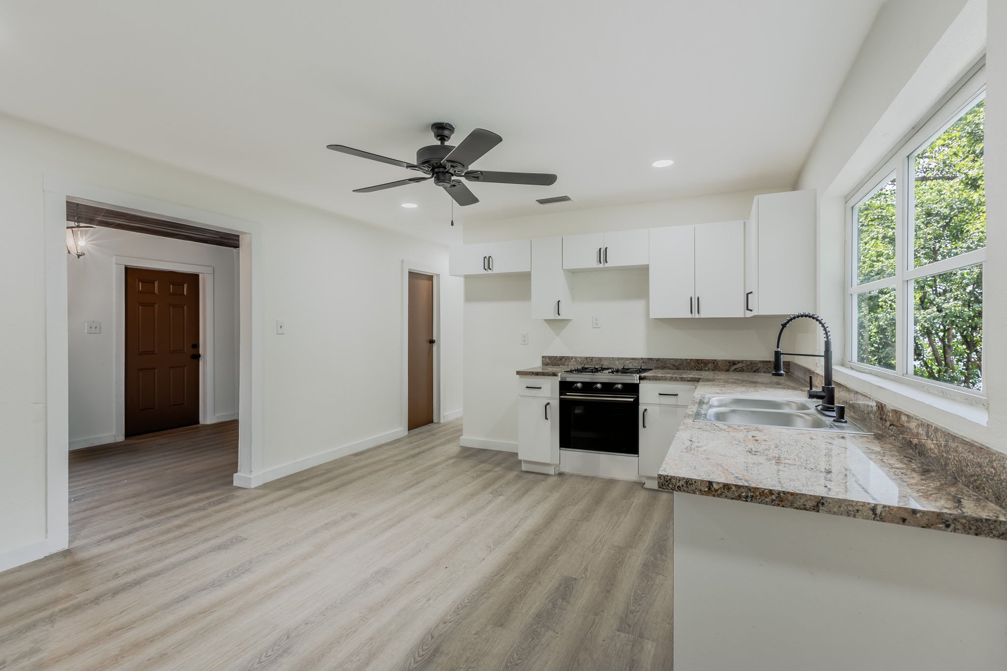 Open kitchen with white cabinets, granite countertops, black faucet, stove, large window showing greenery outside, ceiling fan, and light-colored wood flooring.