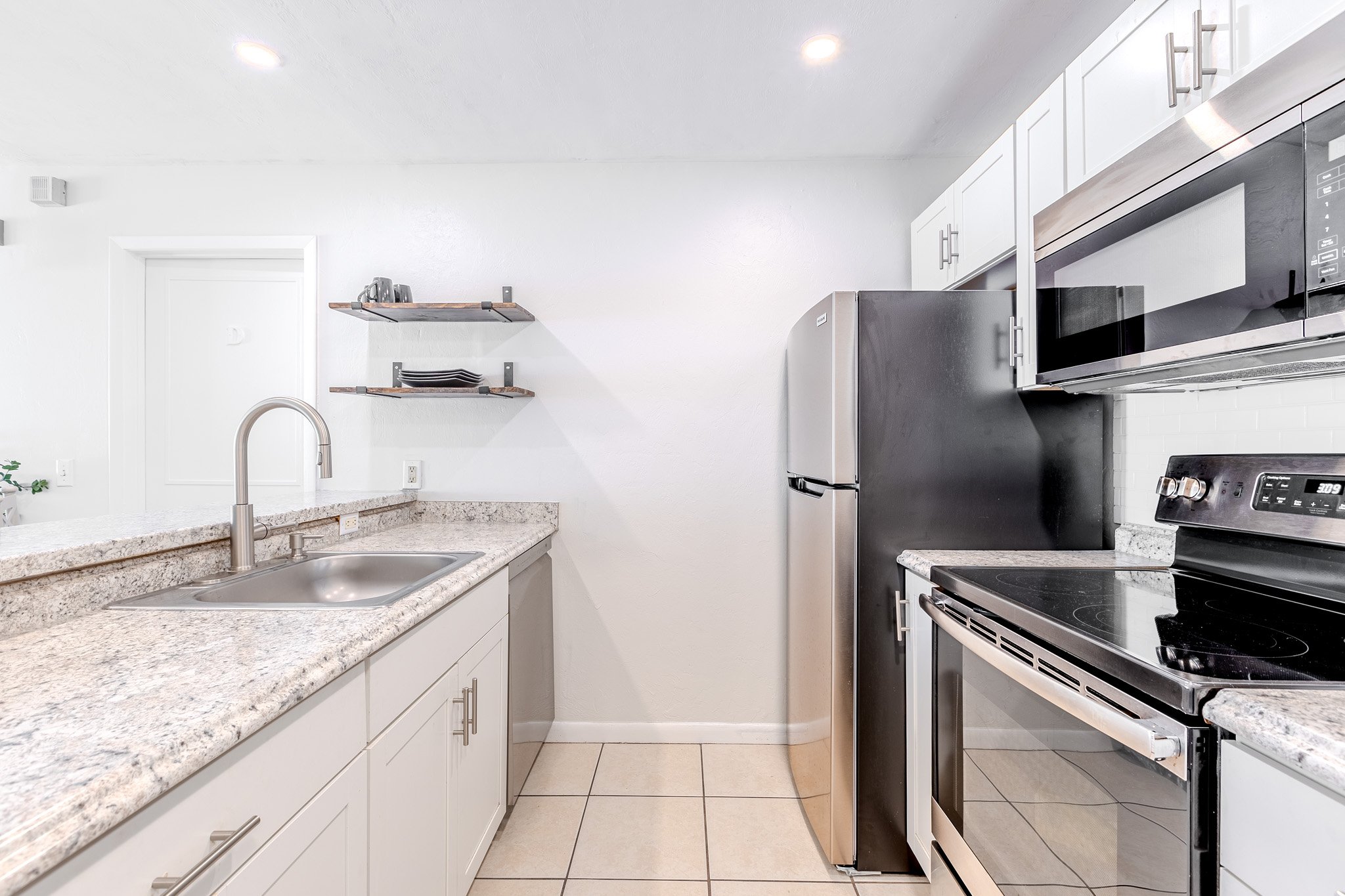 Modern kitchen with white cabinets, granite countertops, stainless steel appliances including a refrigerator, microwave, and oven, and open wall shelves with dishes. Tiled floor and a stainless steel sink.
