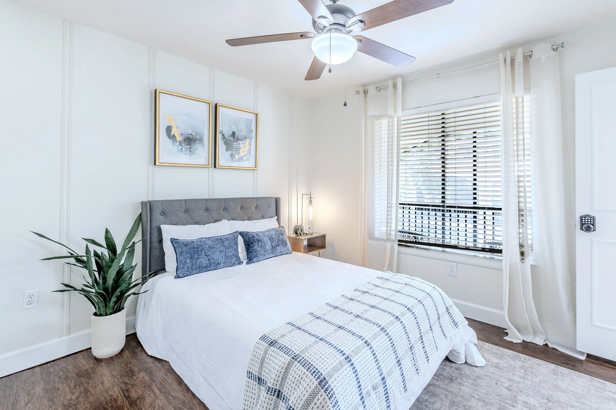 A bedroom with a gray upholstered bed, white bedding, and blue pillows. There are two abstract art prints with gold frames on the wall, a potted plant in a white vase, and a large window with white curtains and blinds. A ceiling fan with wooden blade