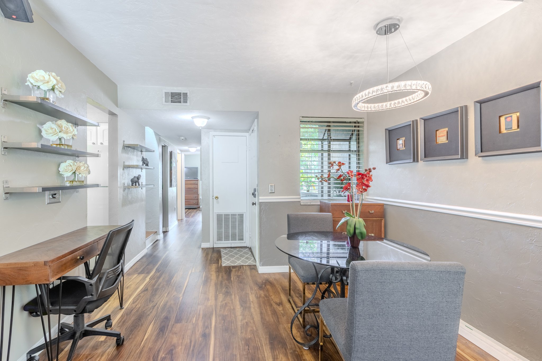 Interior of a living space with a wooden dining table, gray chairs, a round glass coffee table, a floral arrangement in a vase, wall art, window with blinds, and decorative shelves with flowers and animal figurines.