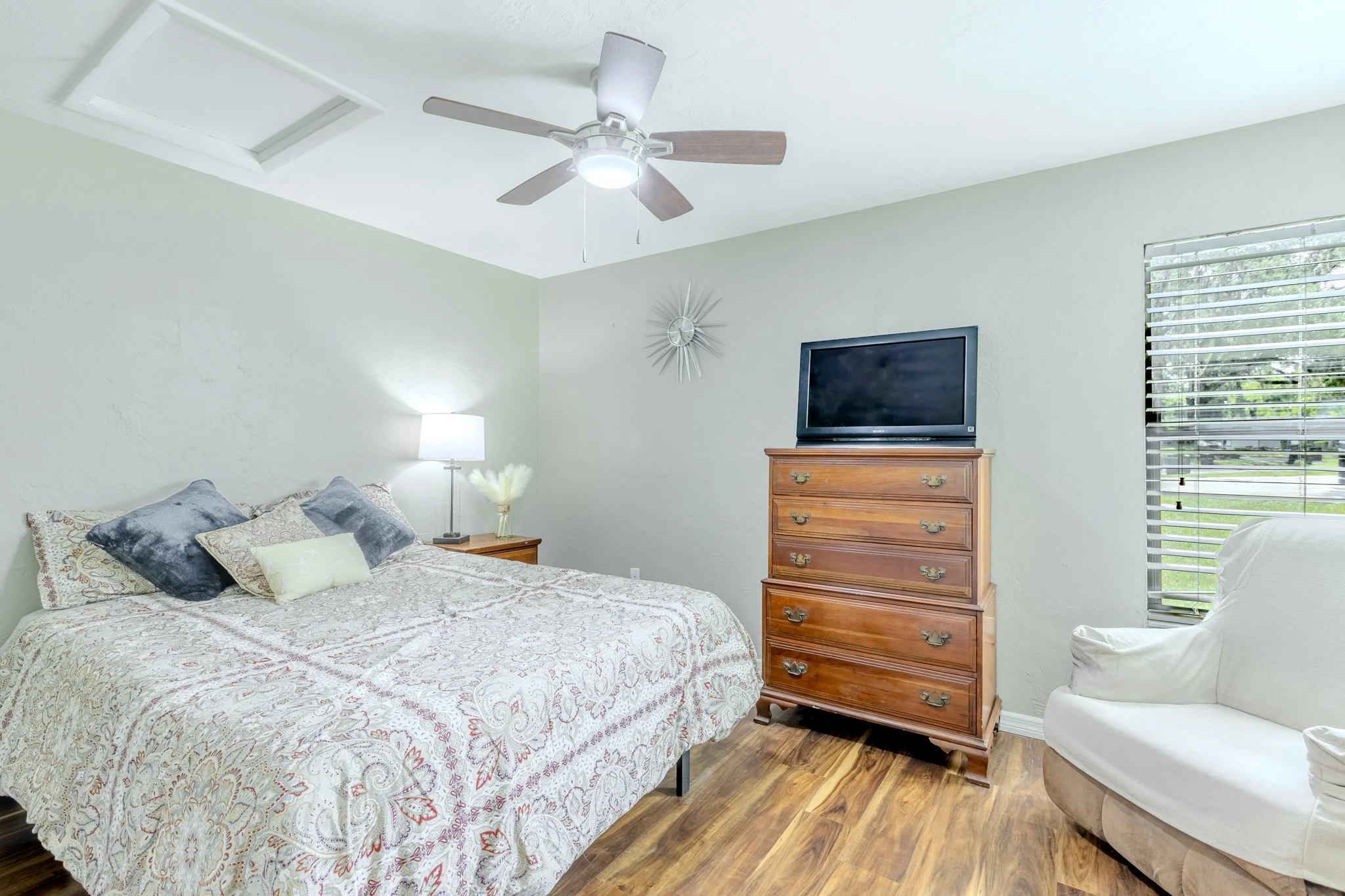 A bedroom with a bed covered in a patterned bedspread and decorative pillows, a wooden dresser with a TV on top, a white armchair, a window with blinds, a wall clock, and a ceiling fan with lights.