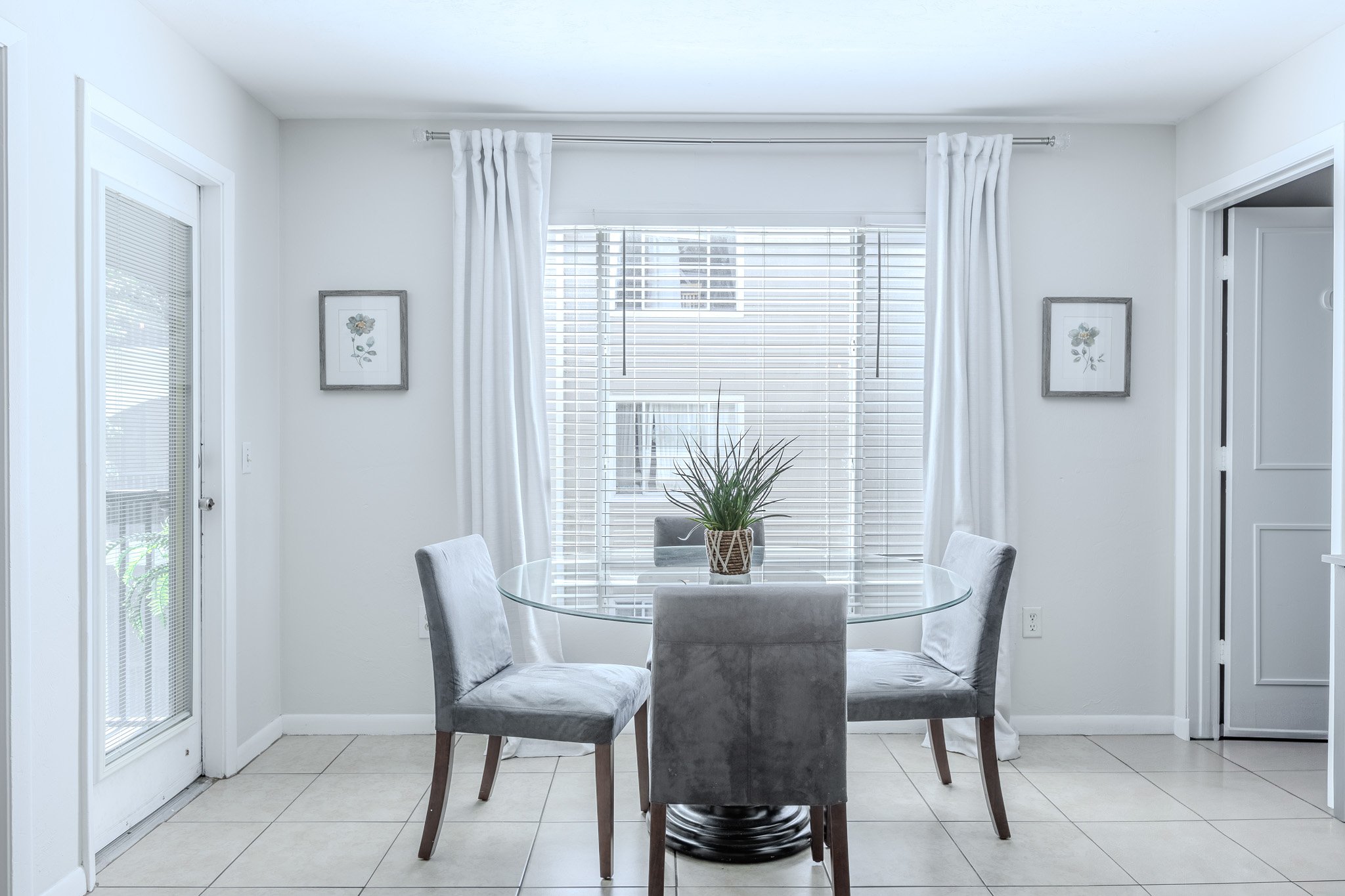 A bright dining area with a round glass table and four gray upholstered chairs. A potted plant sits on the table. The background features large windows with white blinds and curtains, and framed floral artwork on the white walls.
