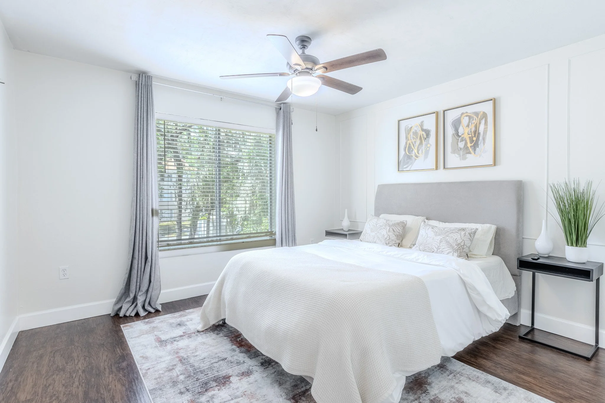 A bright bedroom with a large window with blinds and gray curtains, a ceiling fan with a light, artwork on the wall, a bed with white bedding, a nightstand with a white lamp, and a tall plant in a white vase.