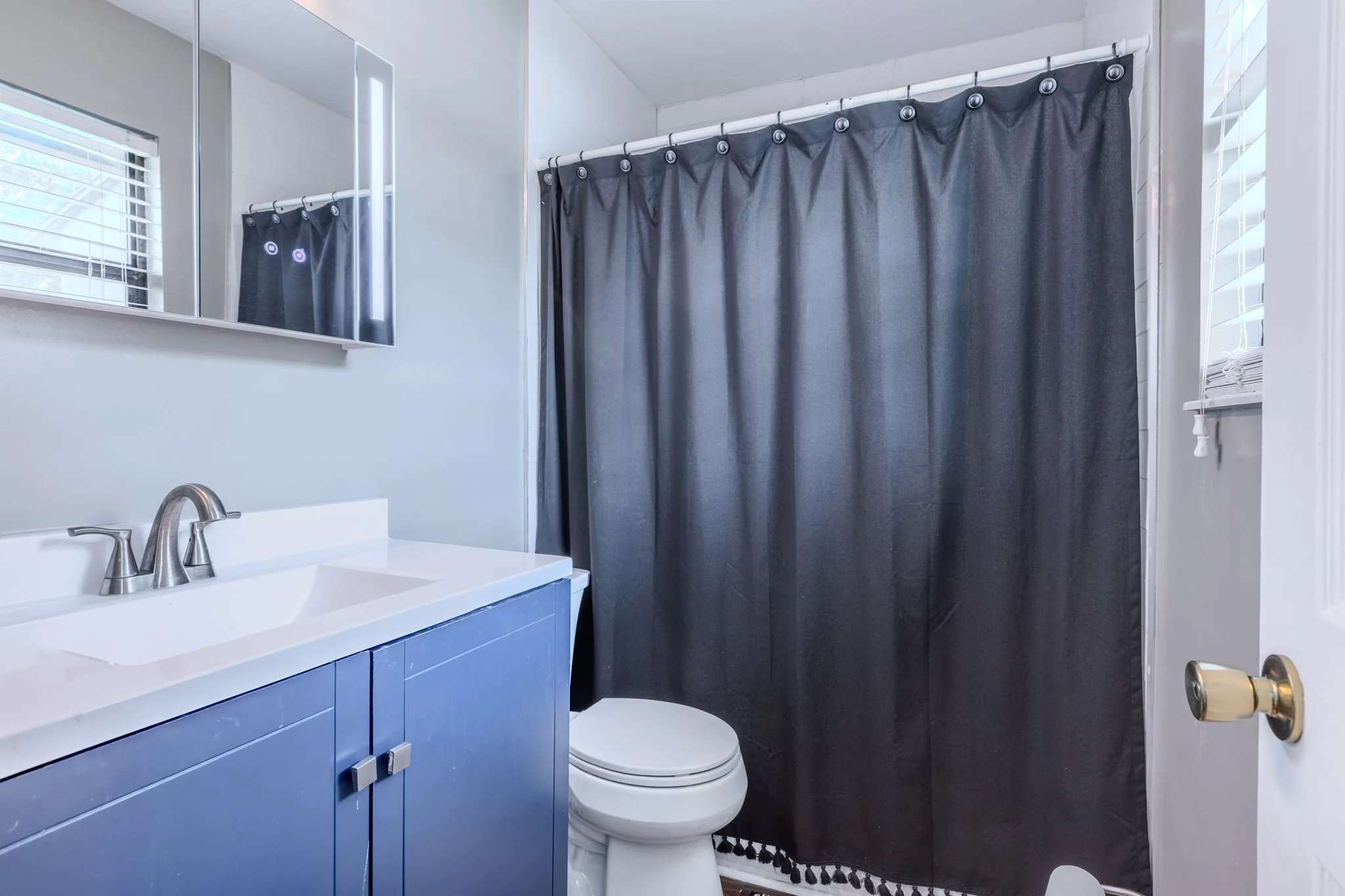 A bathroom with a white sink, blue cabinet, a window with white blinds, a toilet, a mirror cabinet, and a dark shower curtain.