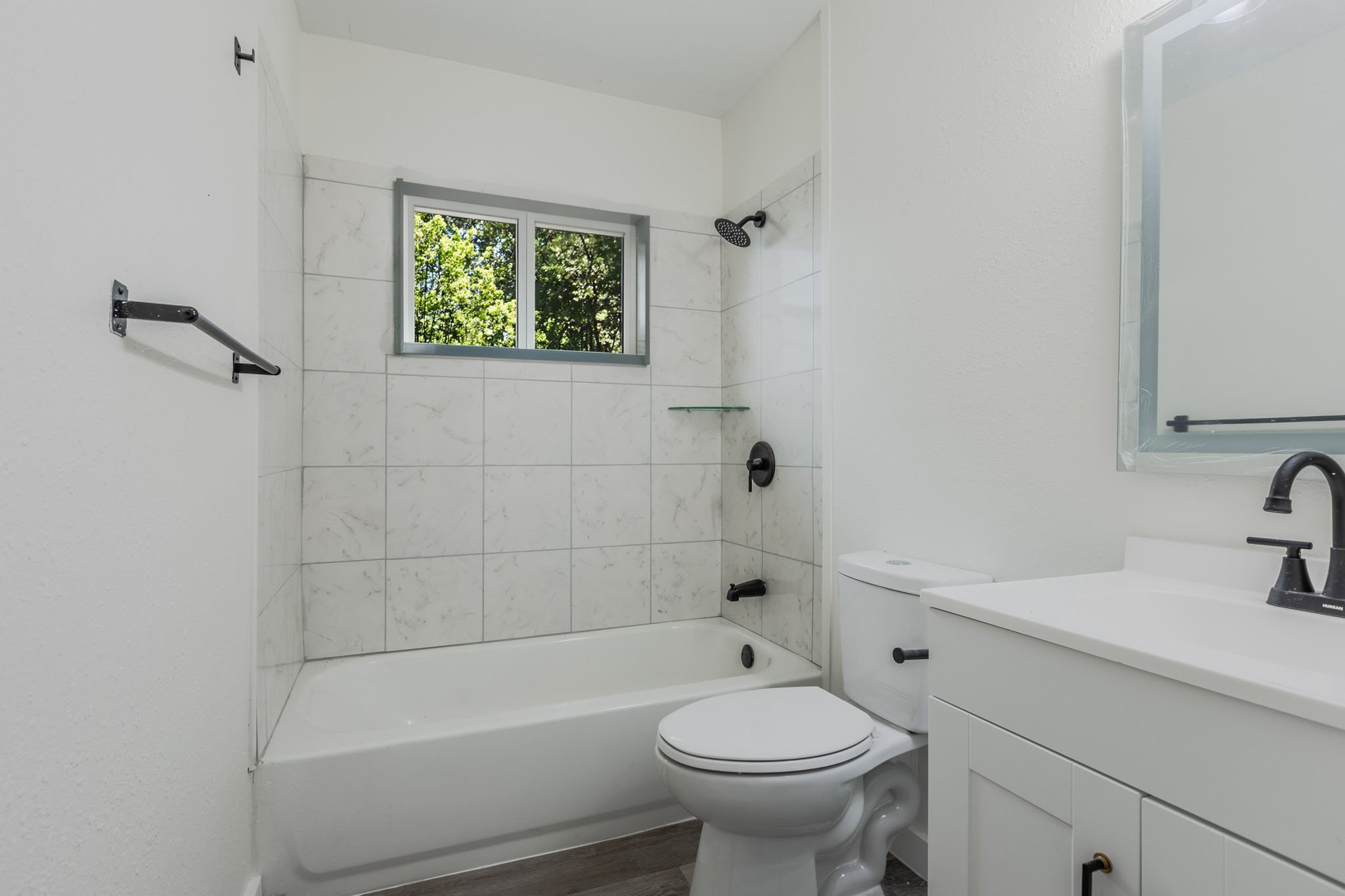 A modern bathroom with a bathtub shower combo, white walls, a frosted window showing greenery outside, a white toilet, a white vanity with black hardware, and a large mirror.