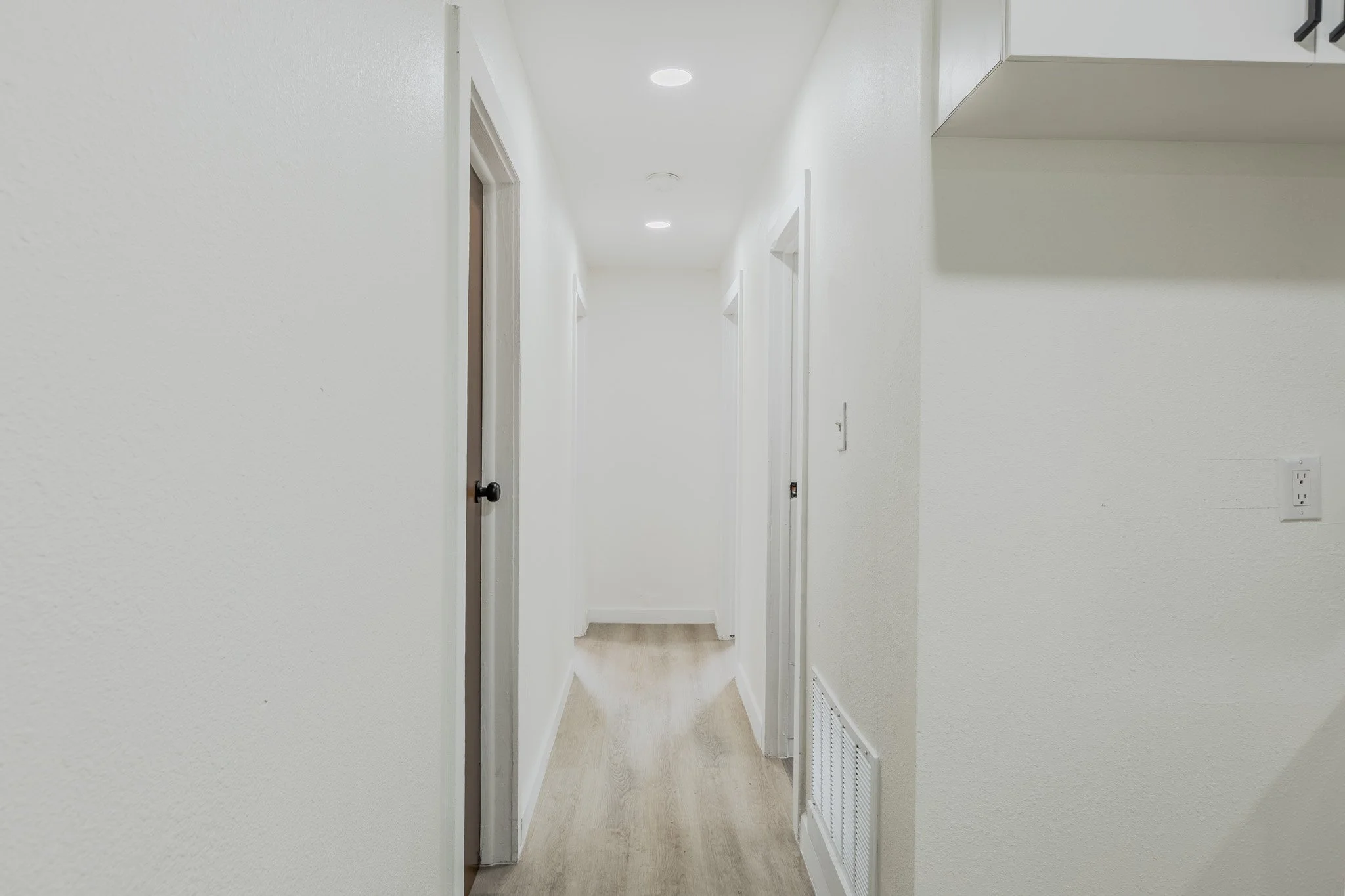 A white hallway with multiple doors on each side, recessed ceiling lights, and a wooden floor.