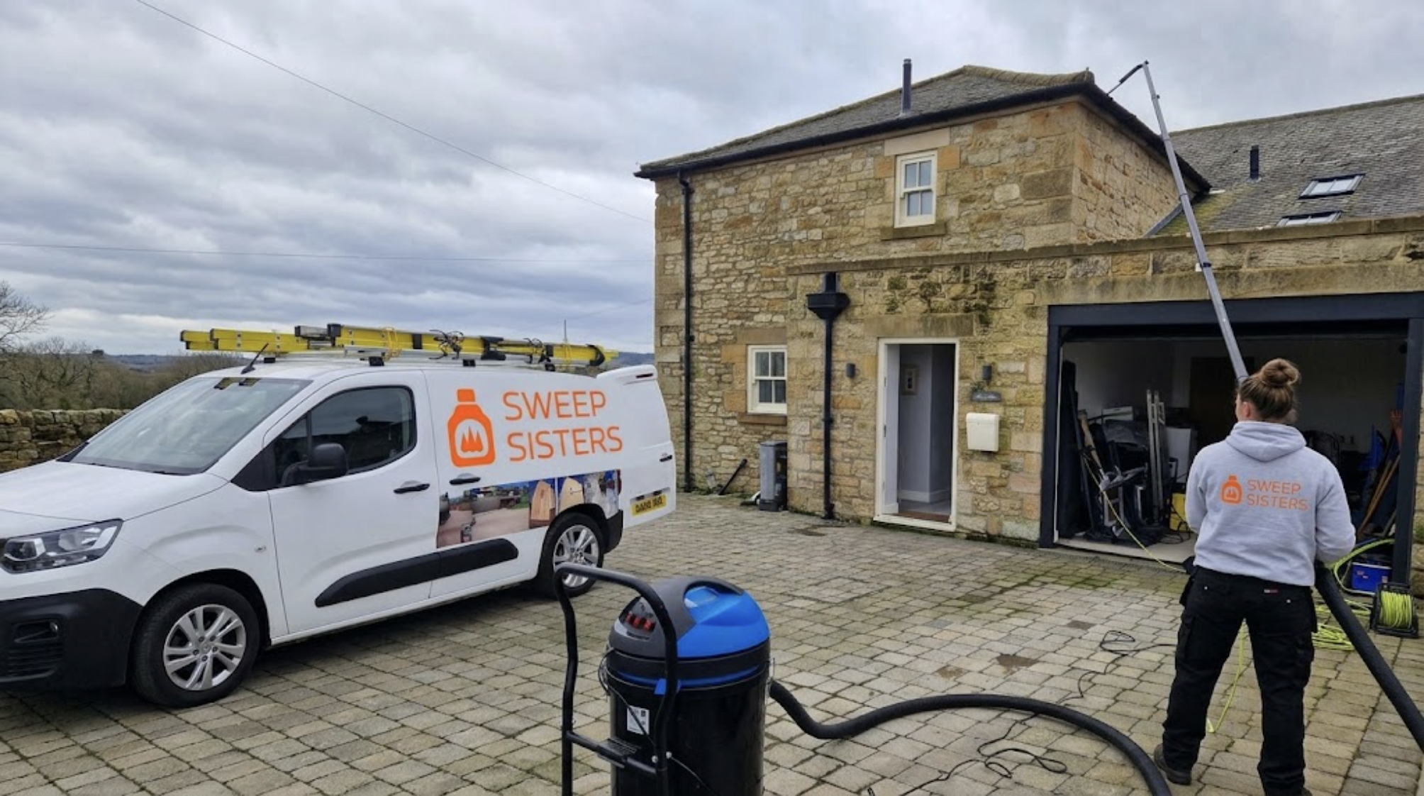 A cleaning service team called Sweep Sisters is washing windows on a house using an extended pole. A white van with the company's logo and name is parked in the driveway, and various cleaning equipment is visible around the area. The house is a stone