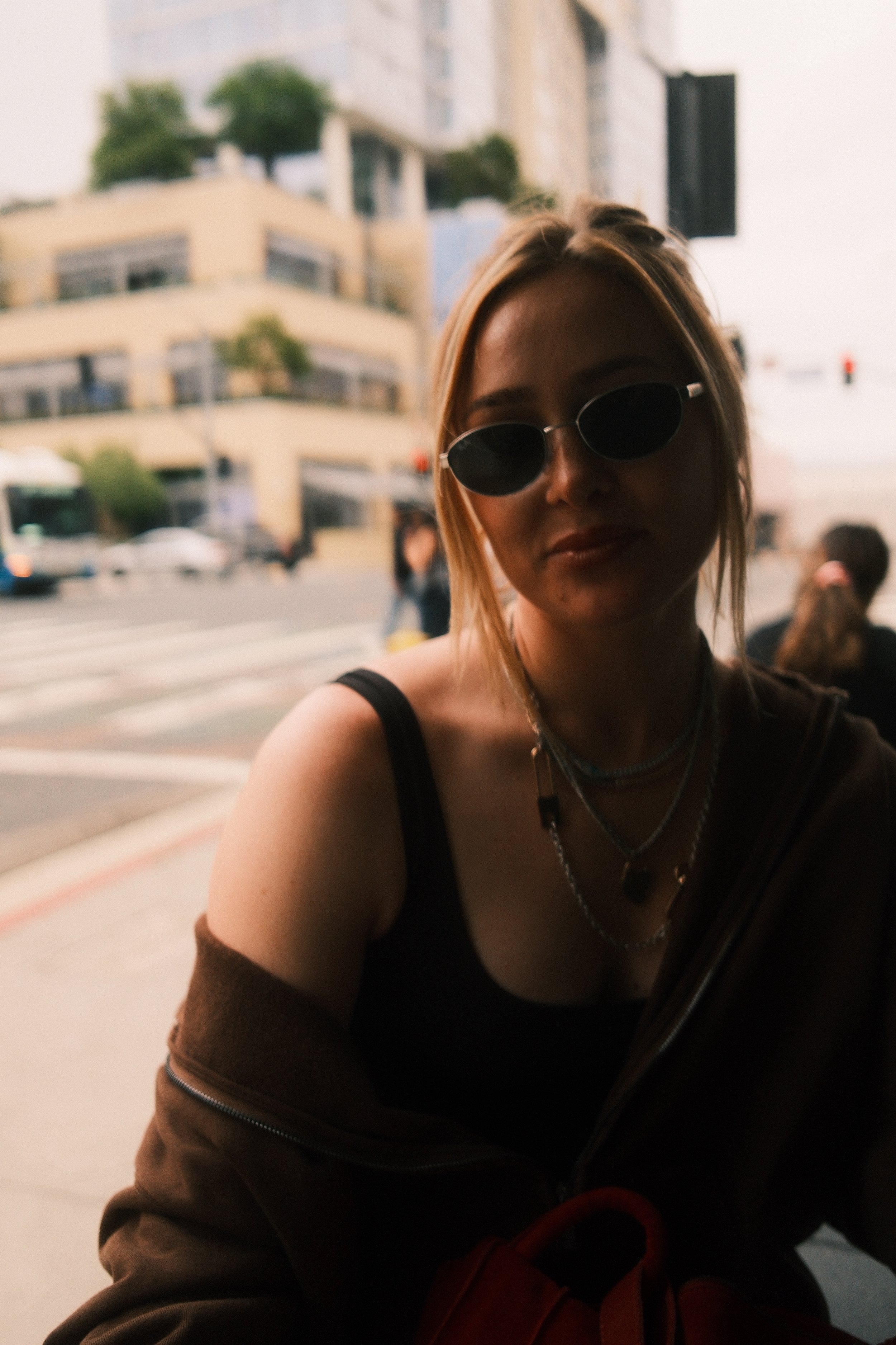 A woman wearing sunglasses and layered necklaces, smiling softly, with a blurred cityscape background that includes multi-story buildings and parked cars.