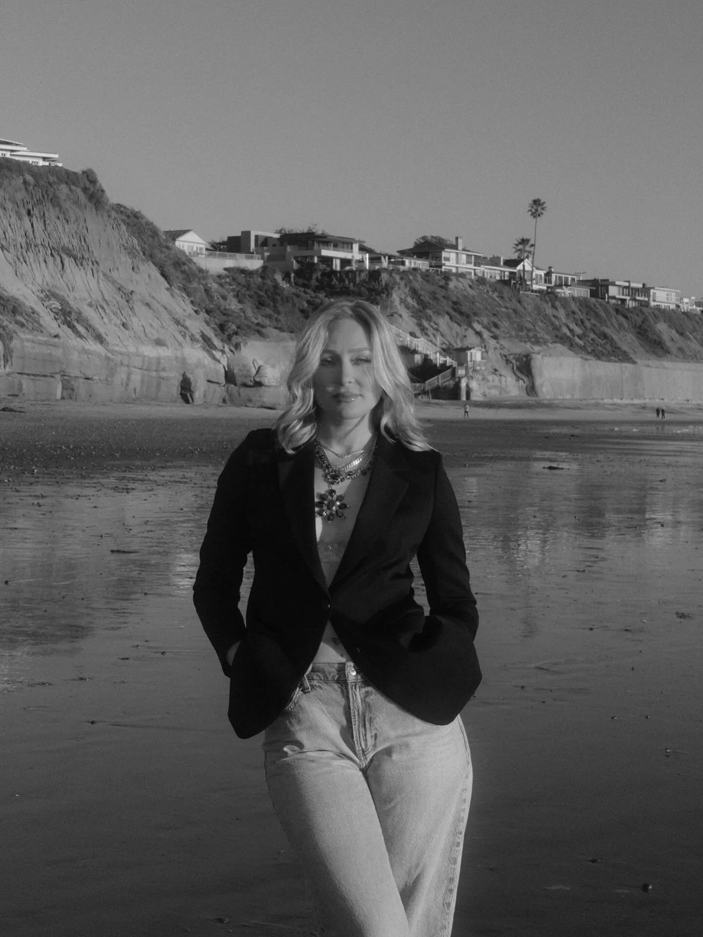 A woman with wavy blonde hair stands on the beach with her hands in her pockets, wearing a black blazer, layered necklaces, and light-colored jeans. In the background, there are rocky cliffs with modern houses and two tall palm trees, under a clear sky.