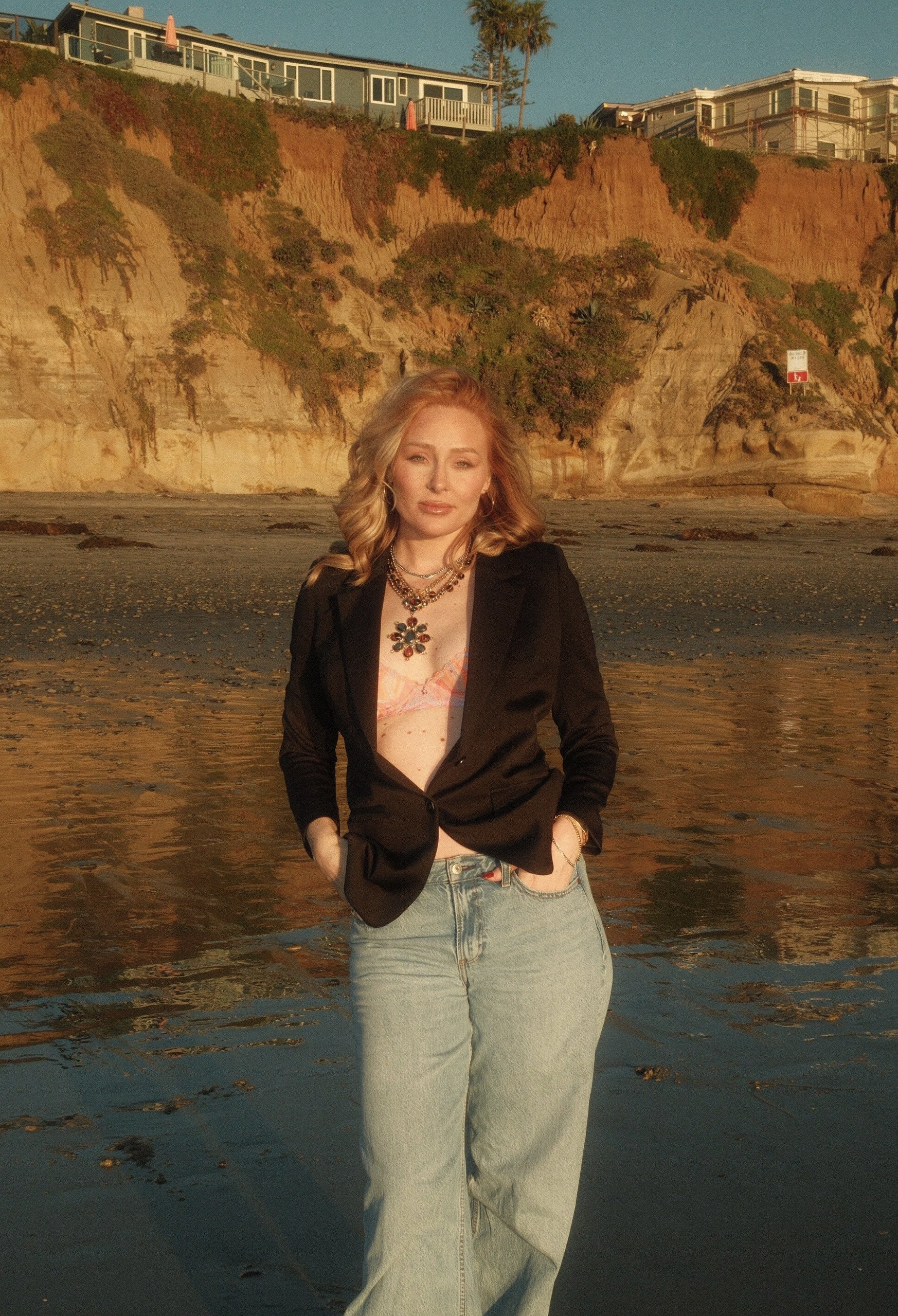 A woman with blonde hair wearing a black blazer, pink top, and light jeans posing on a beach at sunset, with cliffs and houses in the background.