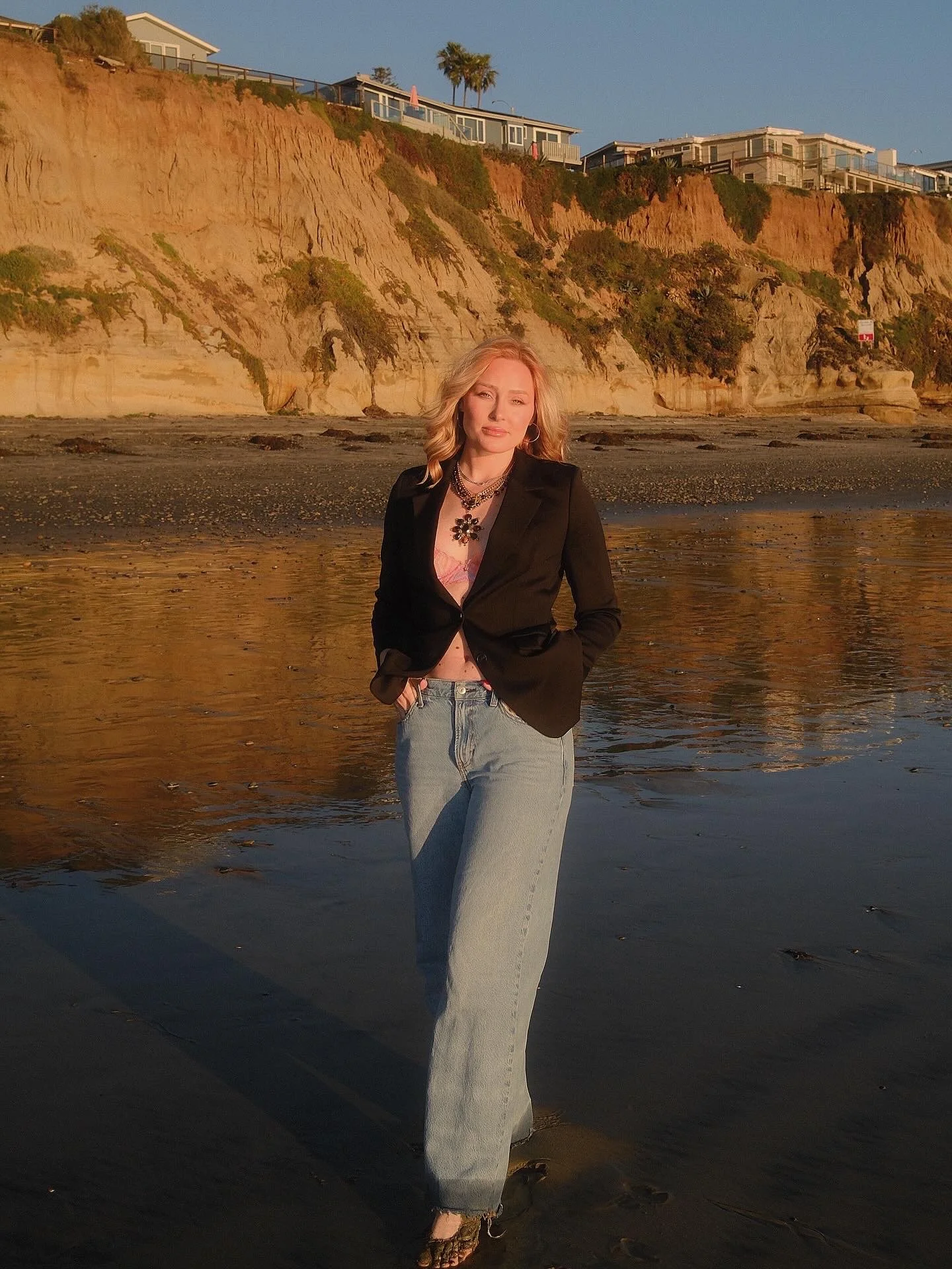 A woman standing in shallow water on a beach during sunset, with cliffs and houses in the background.