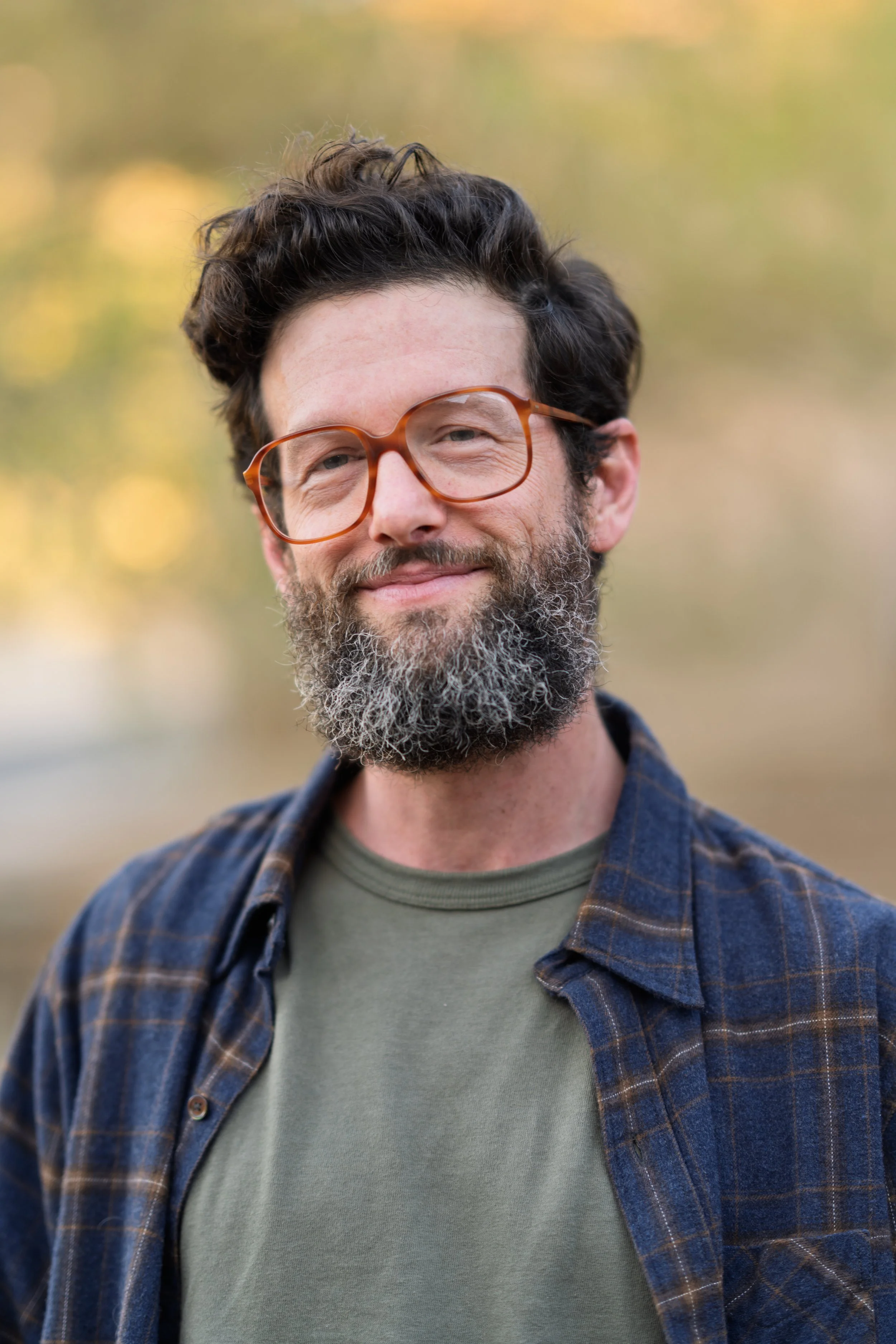 A man with dark, wavy hair, glasses, and a beard, smiling outdoors in front of a blurred background of trees with autumn-colored leaves. He is wearing a gray t-shirt and a blue plaid shirt.