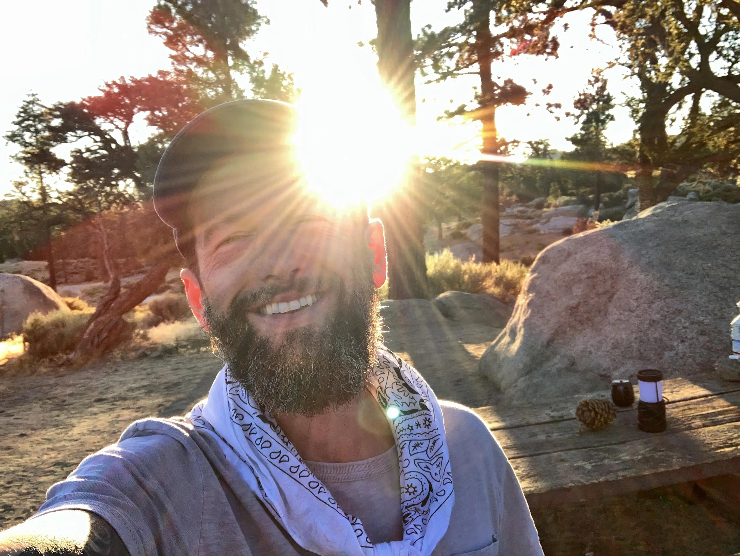 A man with a beard smiling at a camera outdoors during sunset, with trees and large rocks in the background. The sun is shining brightly behind him, creating a lens flare effect.