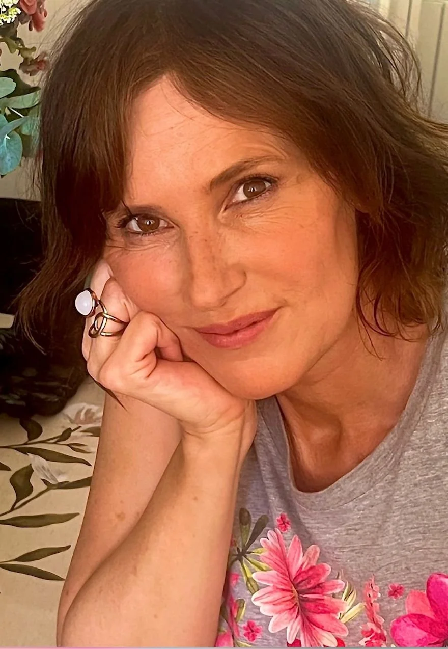 Une femme avec des cheveux bruns, souriante, portant un t-shirt gris avec des motifs floraux roses, posant avec sa tête reposant sur sa main.