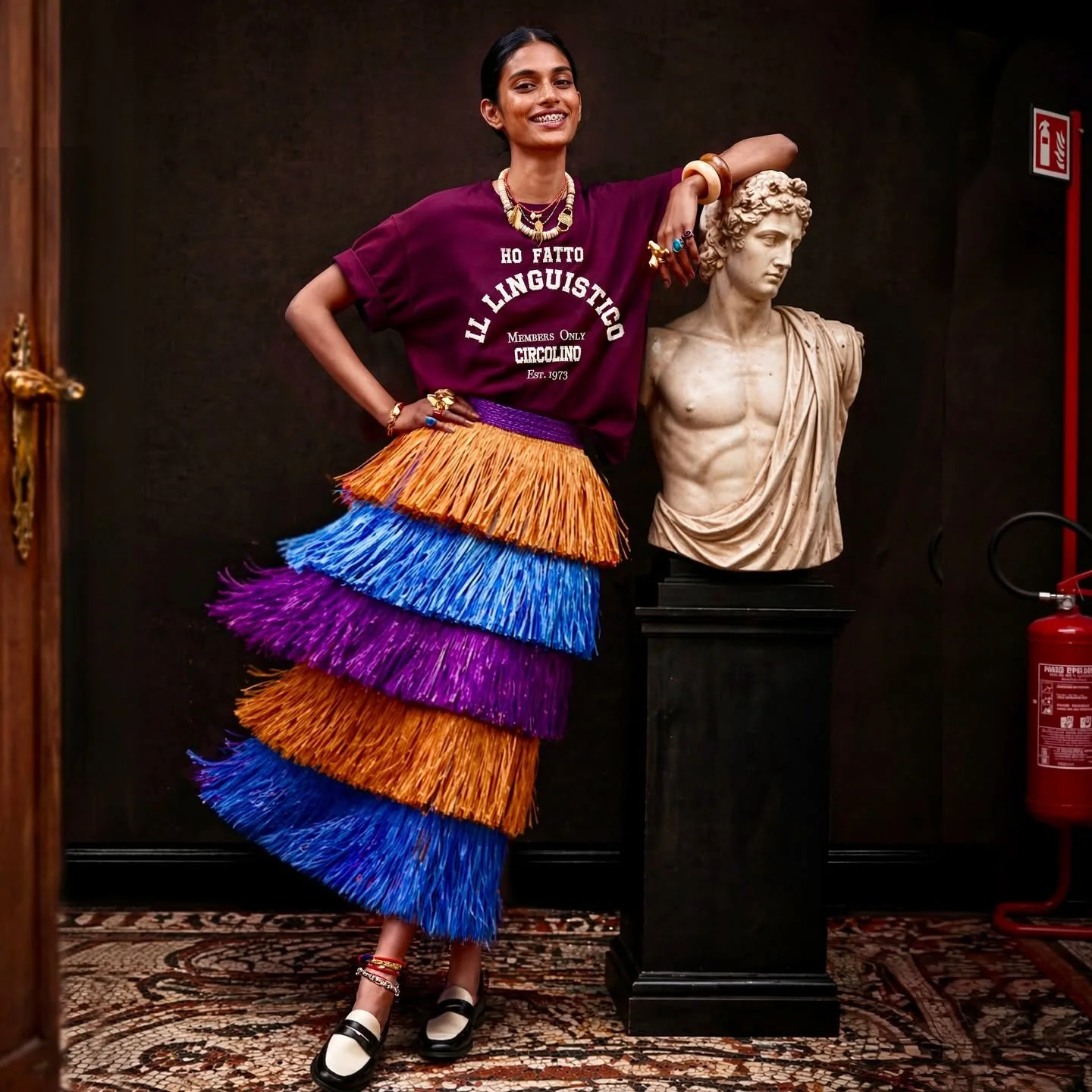 Une femme souriante portant un t-shirt violet avec du texte blanc, une jupe colorée à franges à b r i d d l e, et des chaussures noires et blanches, posant à côté d'une statue en marbre d'un homme antique.