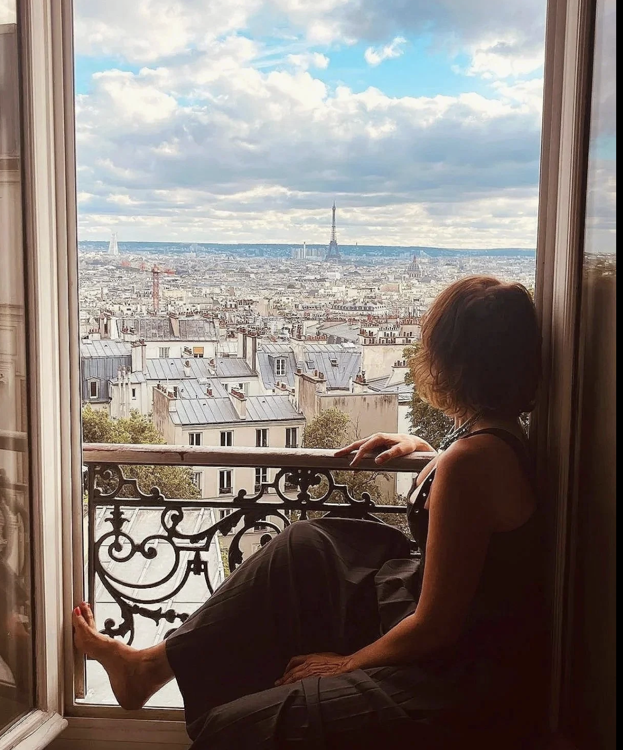 Une femme assise à un balcon avec vue sur la ville de Paris, la Tour Eiffel au centre en arrière-plan, le ciel bleu avec quelques nuages.