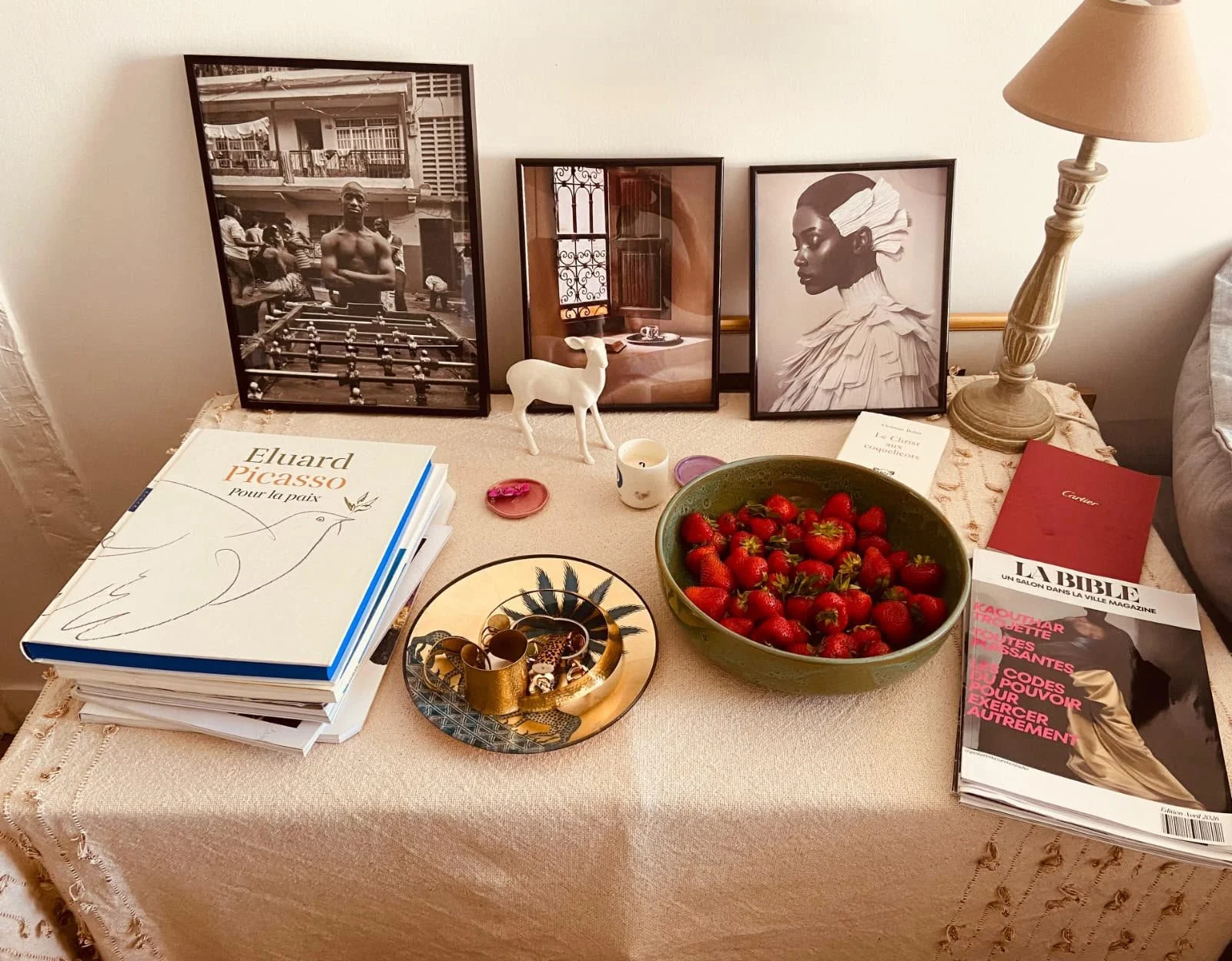 Tableau avec des livres, une coupe de fraises, une petite figurine de chevreuil blanche, une lampe, et des cadres de photos en arrière-plan.