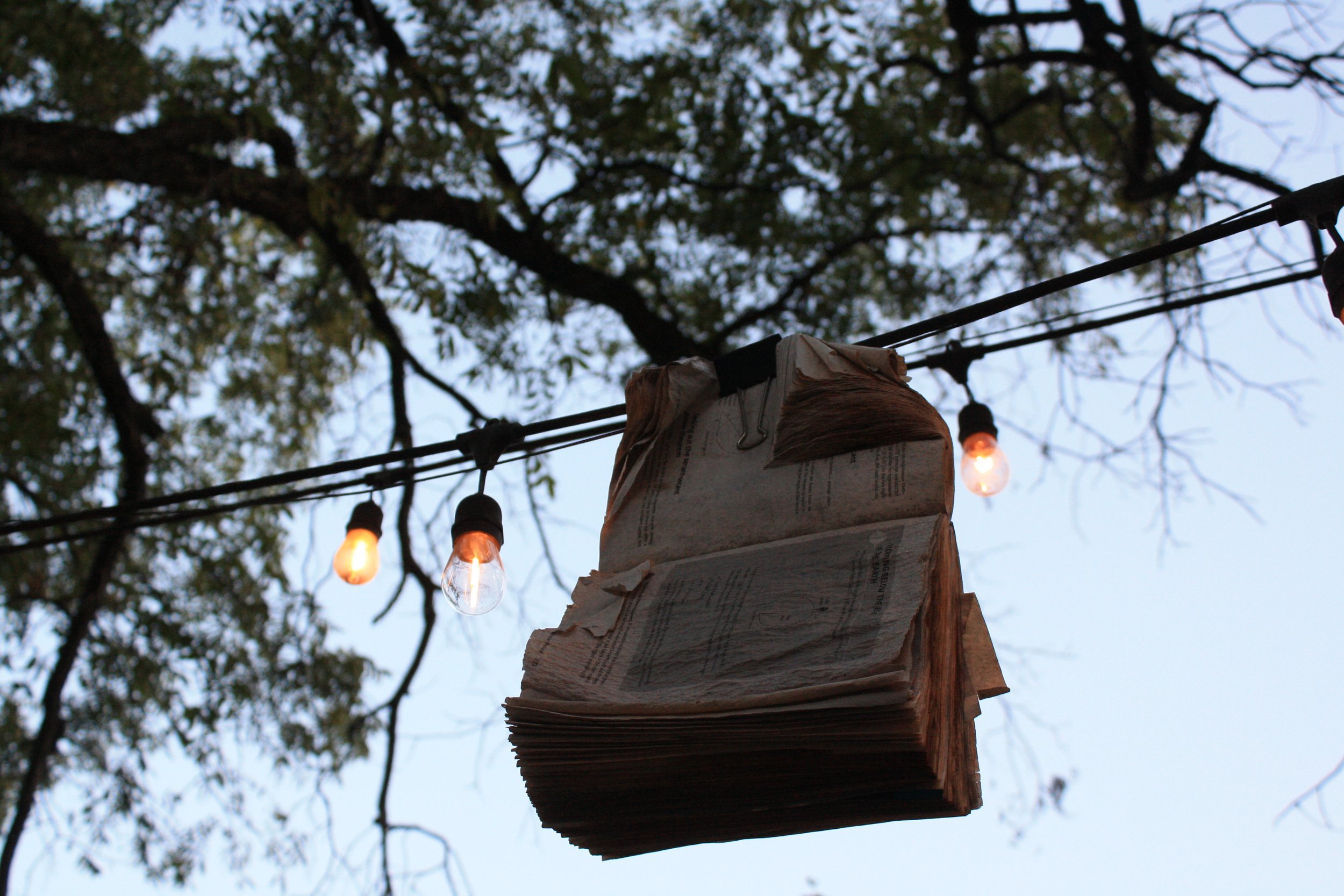A makeshift hanging lamp made of paper and newspaper attached to a wooden post with string, illuminated by small Edison-style light bulbs, against the backdrop of tree branches and sky.