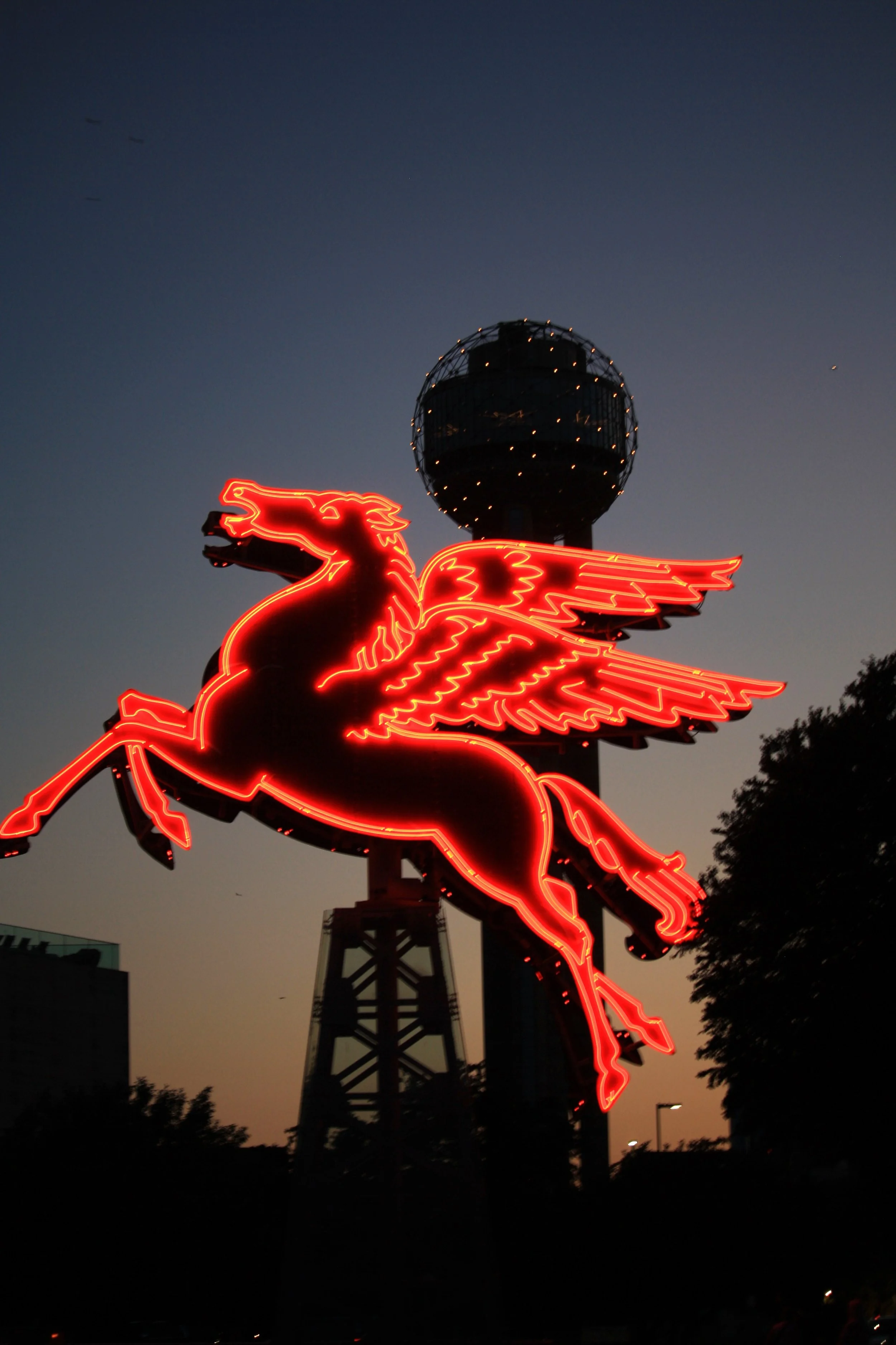 A neon sign of a red Dallas, Texas Pegasus with wings spread, mounted on a structure with an Reunion tower in the background during dusk or nighttime.