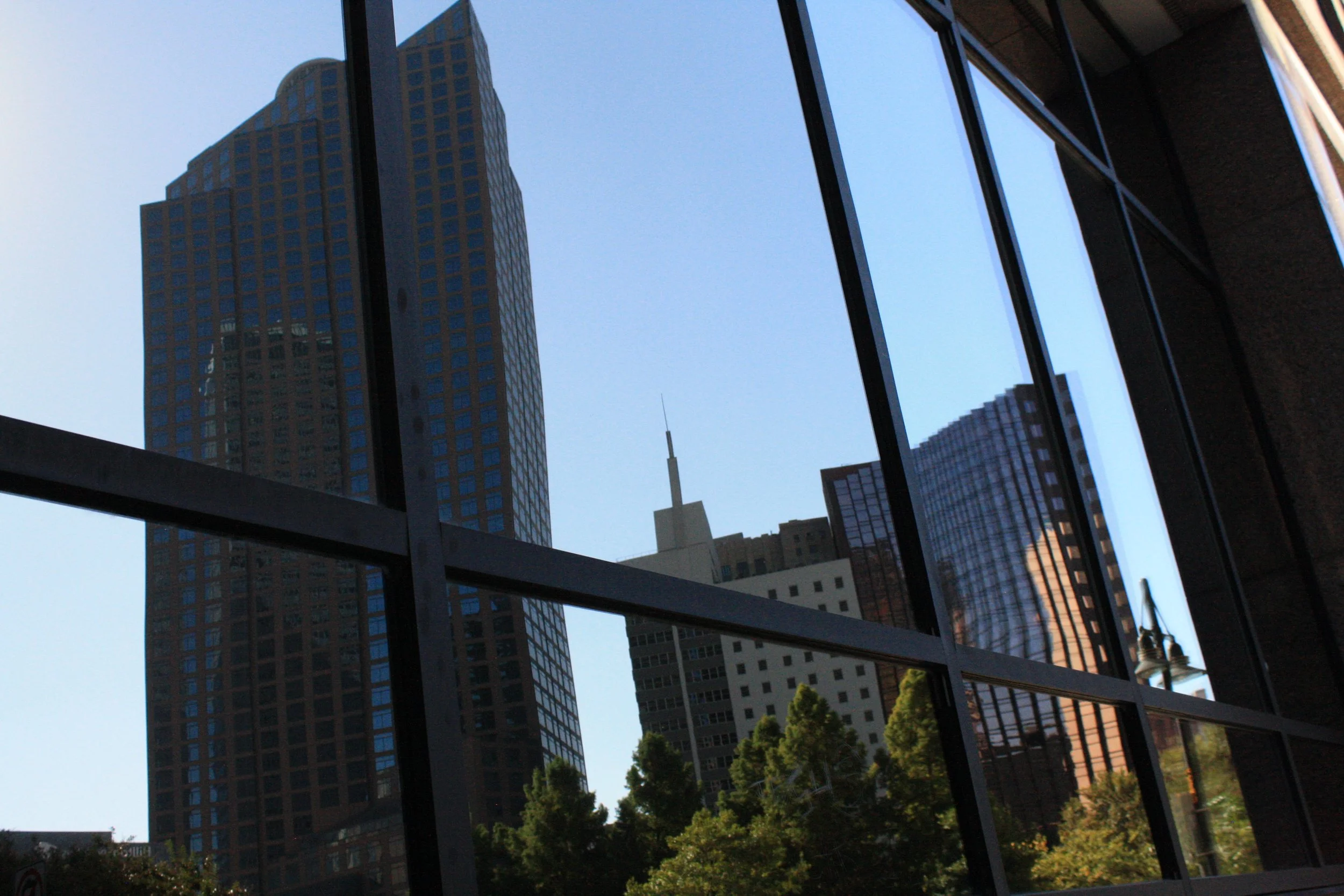 View through a window showing tall skyscrapers and buildings in an urban cityscape with some trees at the bottom.