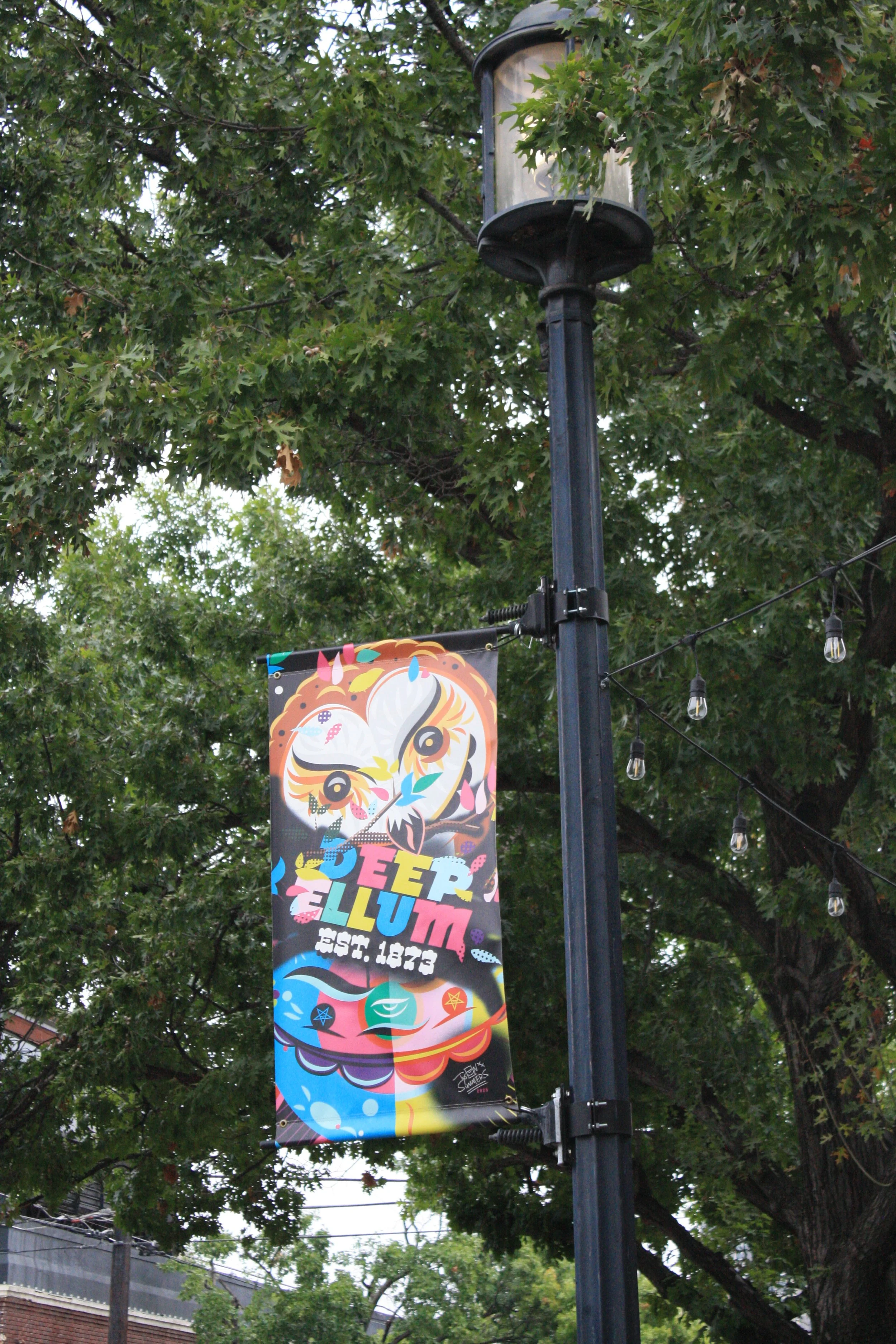 Colorful festival banner hanging from a street lamp post, surrounded by green leafy trees, with string lights and a building in the background.