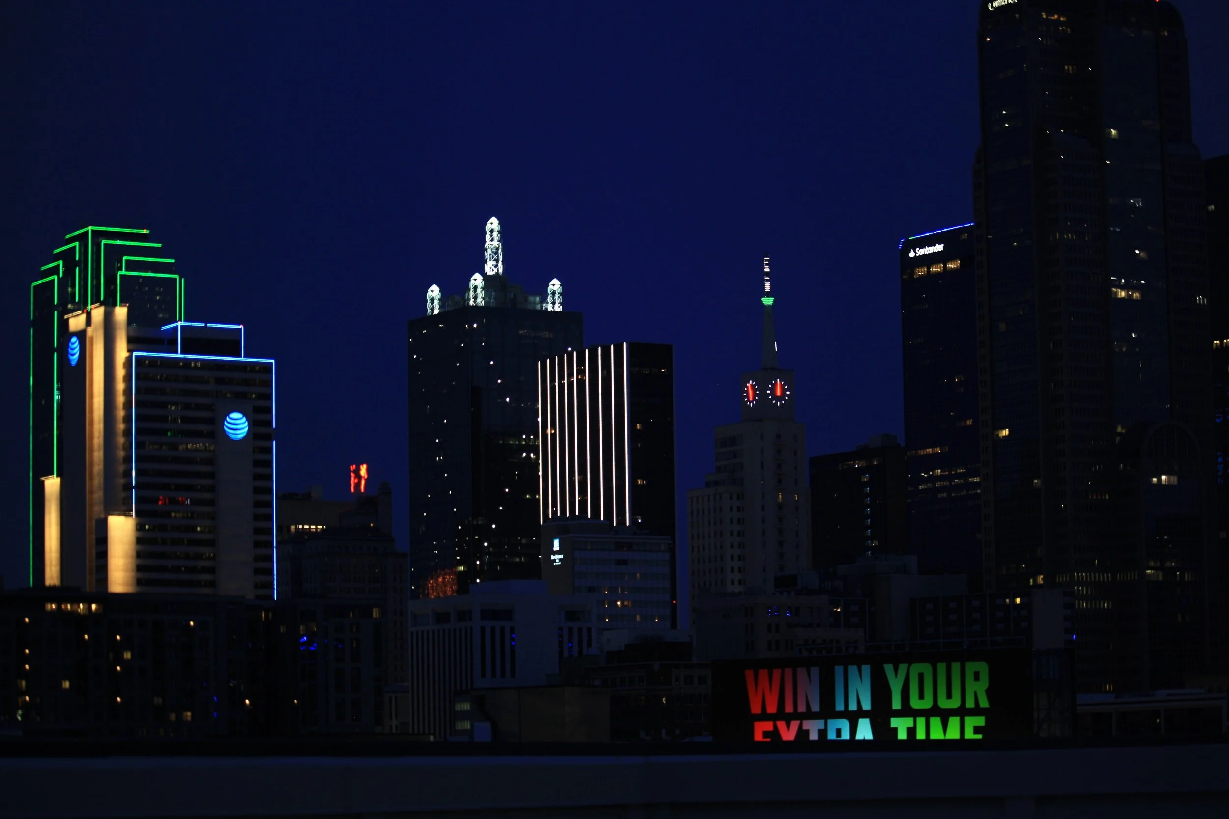 Nighttime Dallas, Texas skyline with illuminated skyscrapers, including a building with green and blue neon outlines, and a tall tower with a green top.
