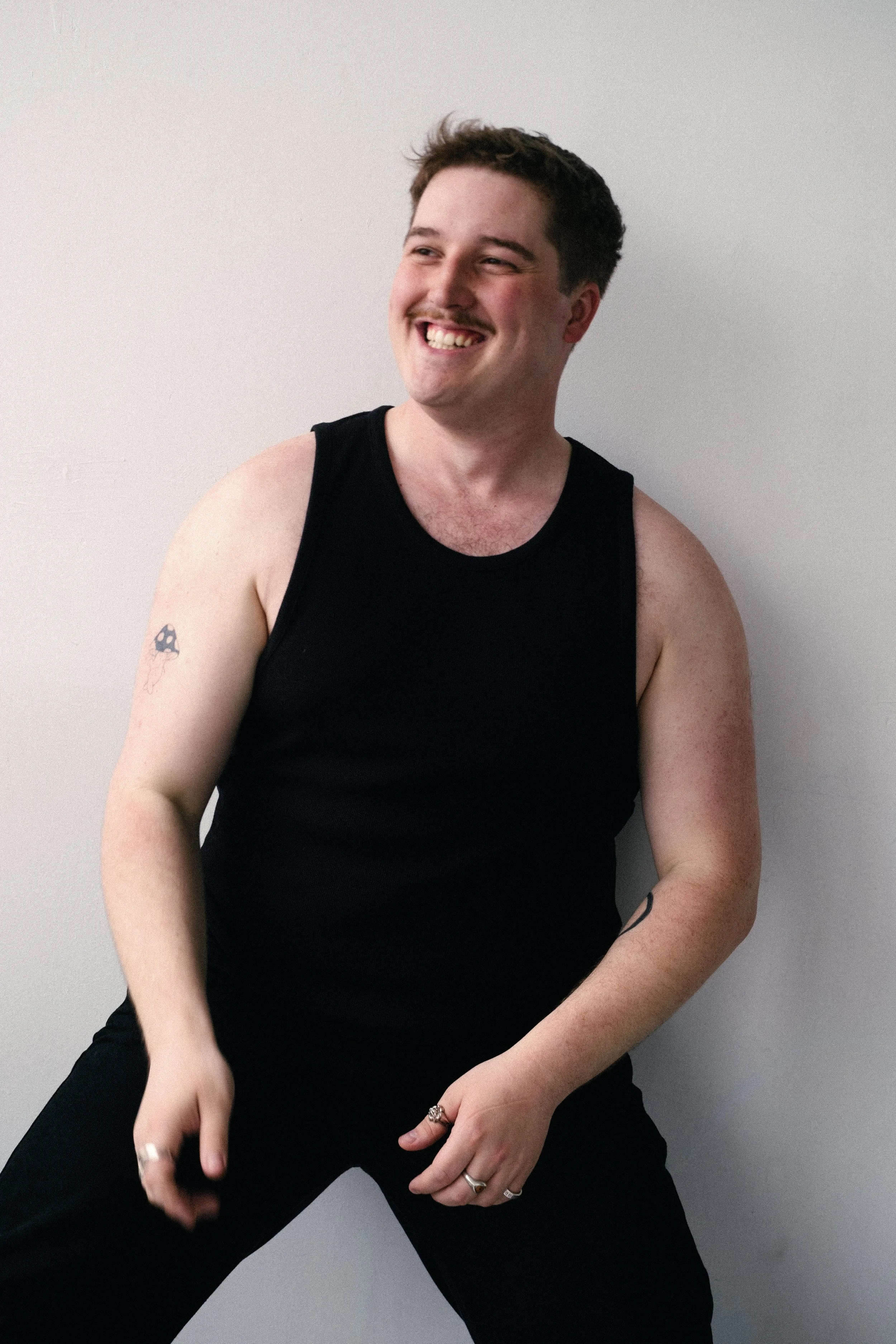 A smiling young man with short dark hair, wearing a black tank top, black pants, and rings on his fingers, standing against a plain white wall.