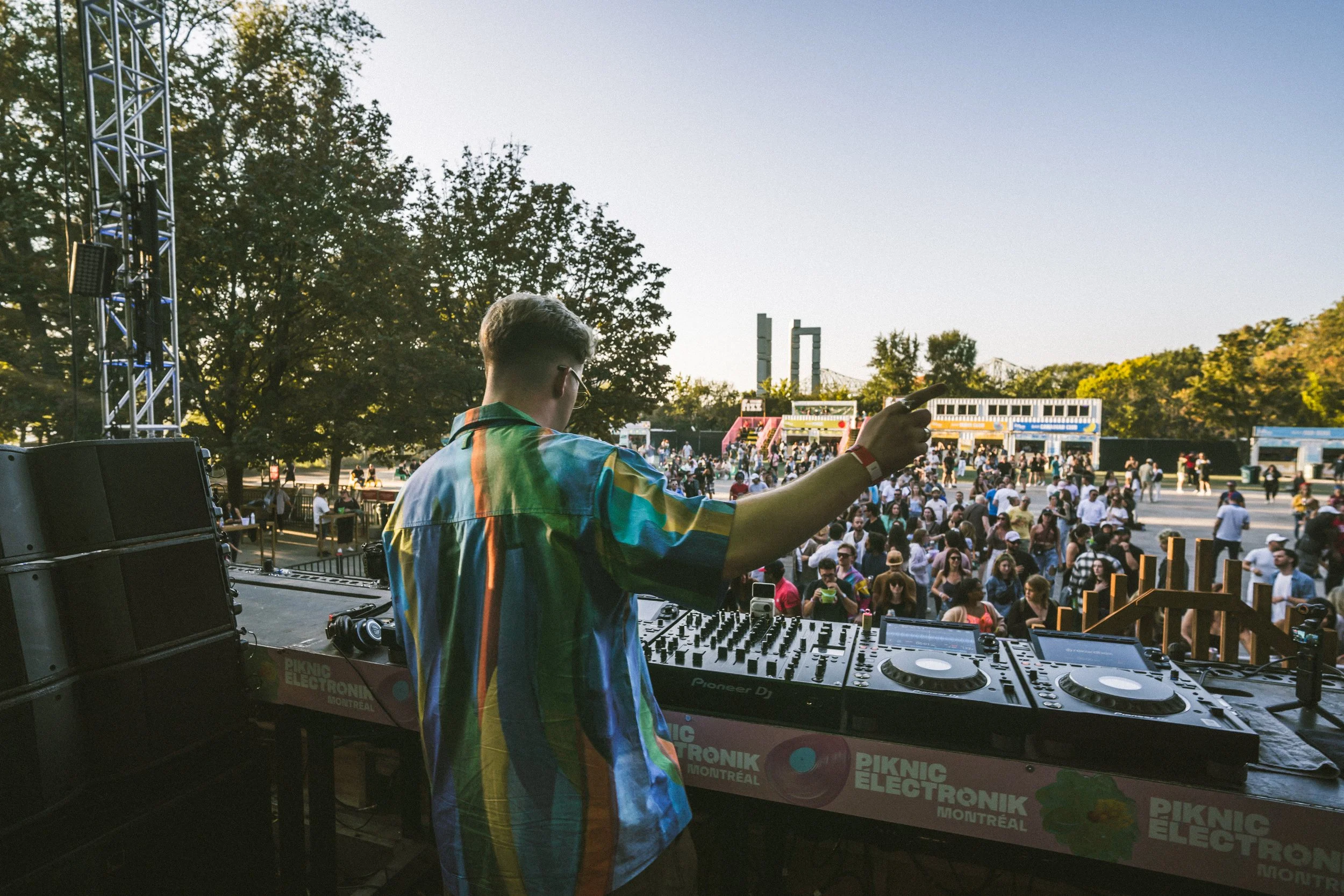 A DJ in a rainbow-colored shirt playing music at an outdoor event with a large crowd, trees, and a bridge in the background during the daytime.