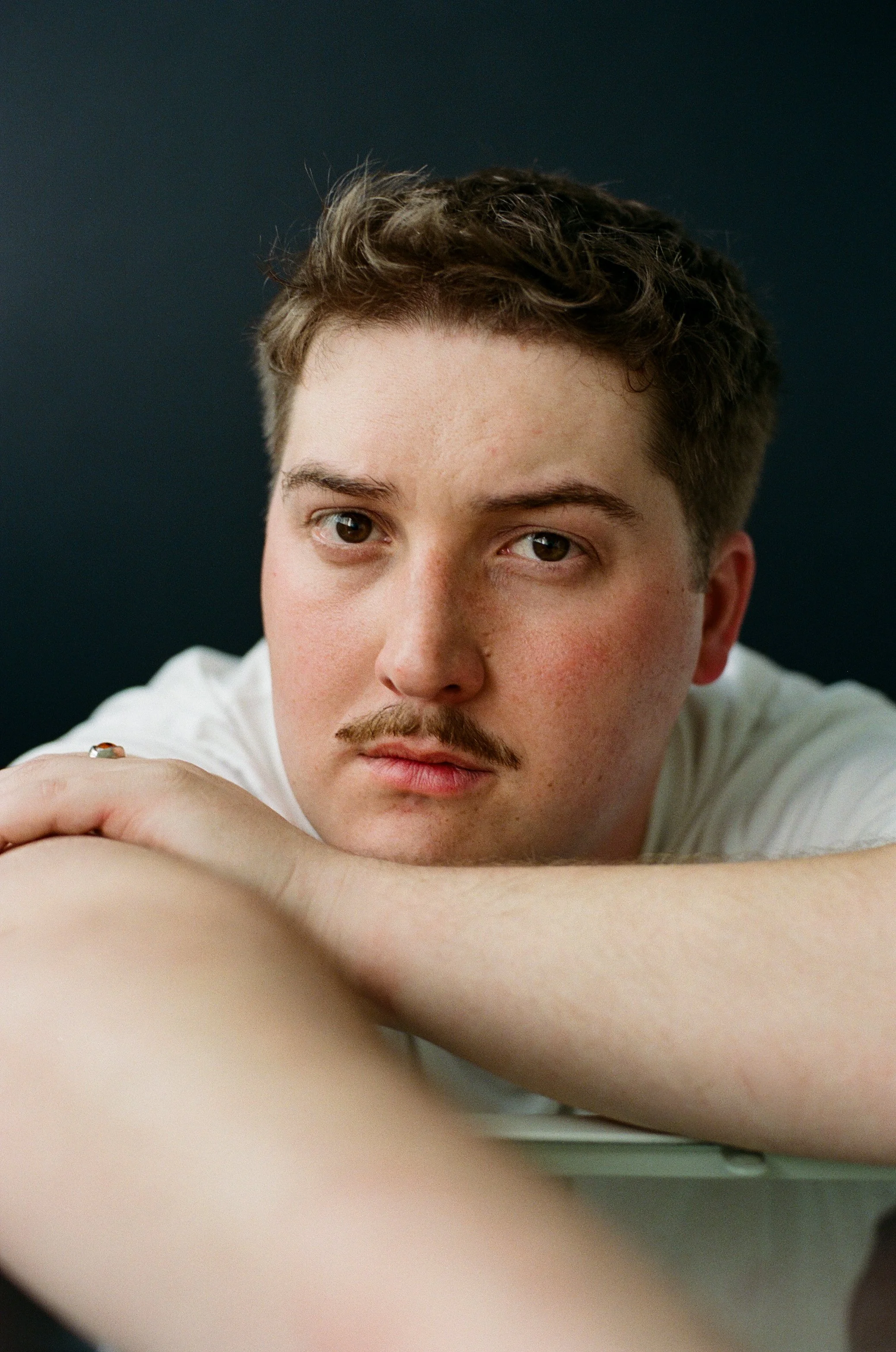 Close-up of a young man with short brown hair and facial hair, resting his chin on crossed arms, looking at the camera with a neutral expression against a dark background.