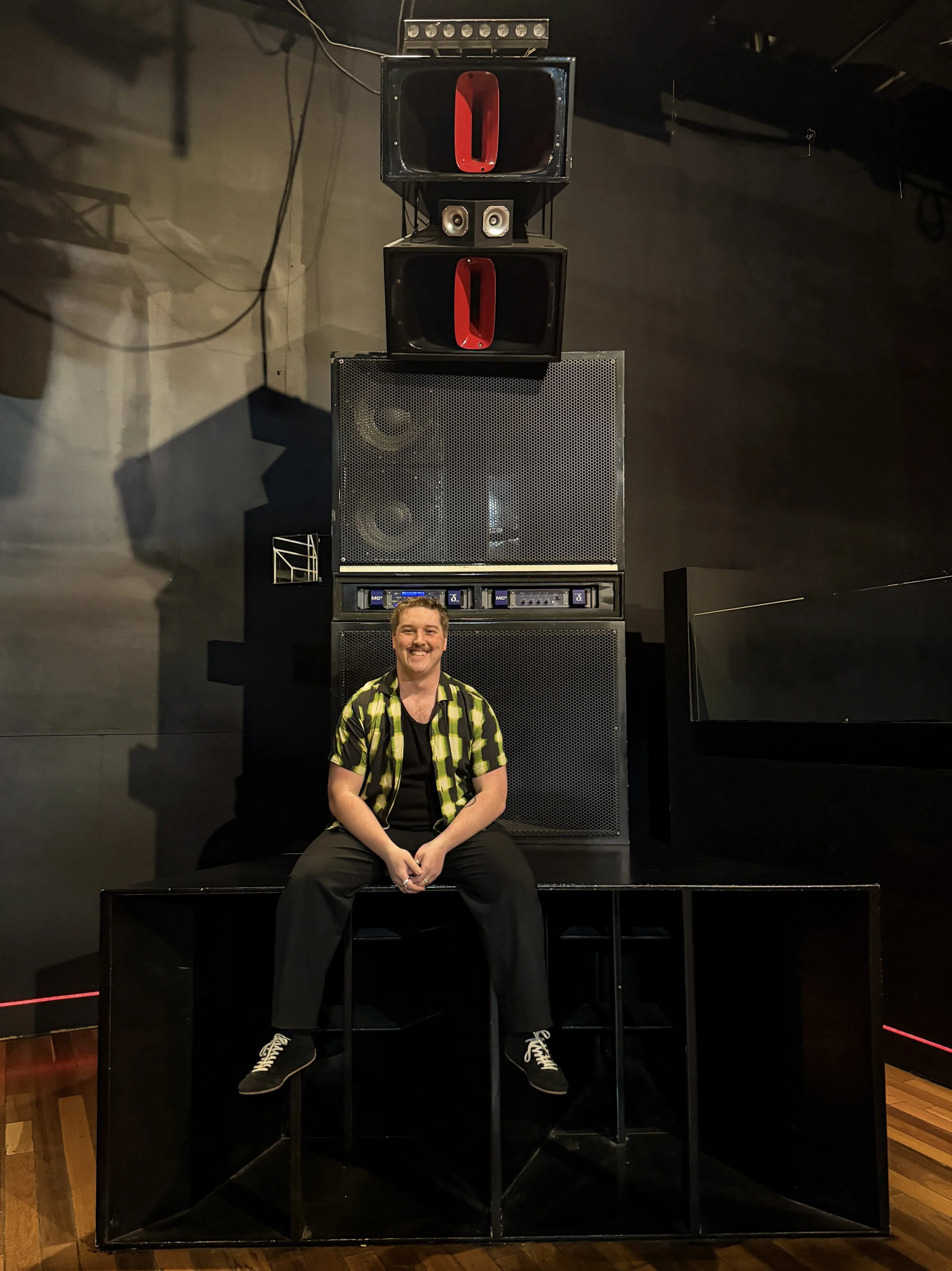 A man with curly hair and a mustache sitting on a black box in front of a large speaker system. The speaker system has two large black speakers with red interiors, a smaller pair of silver speakers, and a rack of audio equipment below. The man is smiling, wearing a black t-shirt, black pants, and a brightly patterned yellow and black shirt.