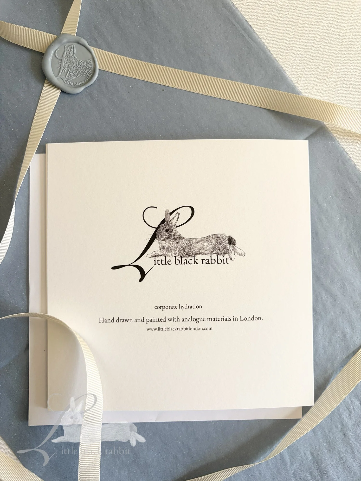 Greetings card back showing the Little Black Rabbit logo, sitting on a white linen tablecloth with antique blue tissue paper, cream ribbon and a blue wax seal.