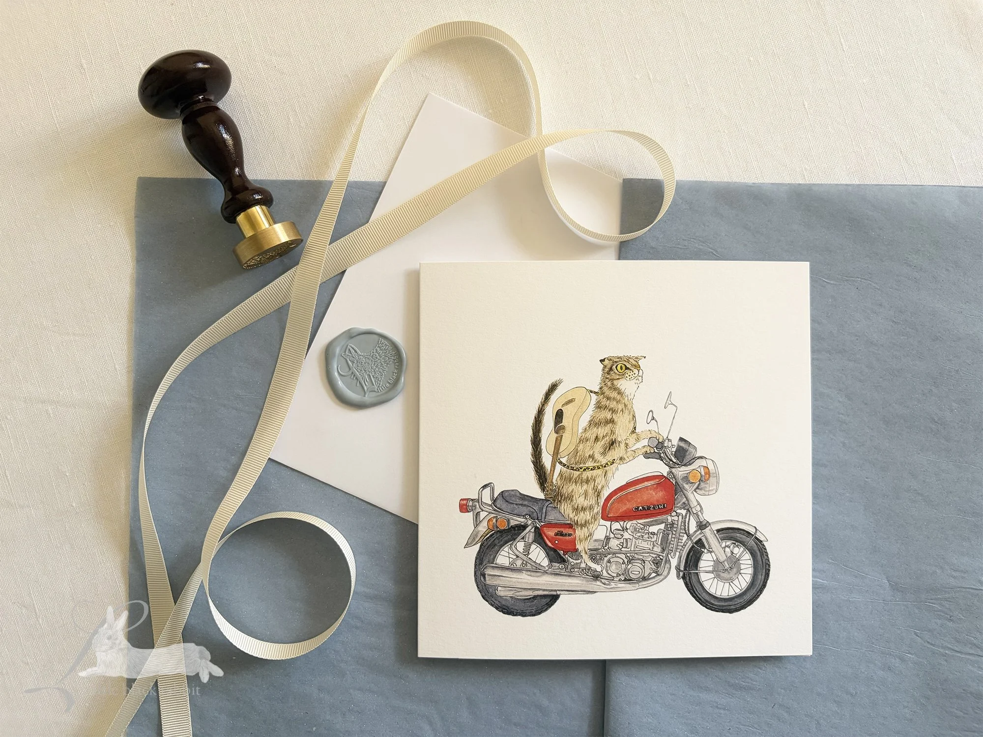 Hand painted greetings card showing a tabby cat on a red motorbike with a guitar on his back, sitting on a white linen tablecloth with cream ribbon, blue tissue paper, and a wax seal with stamp.