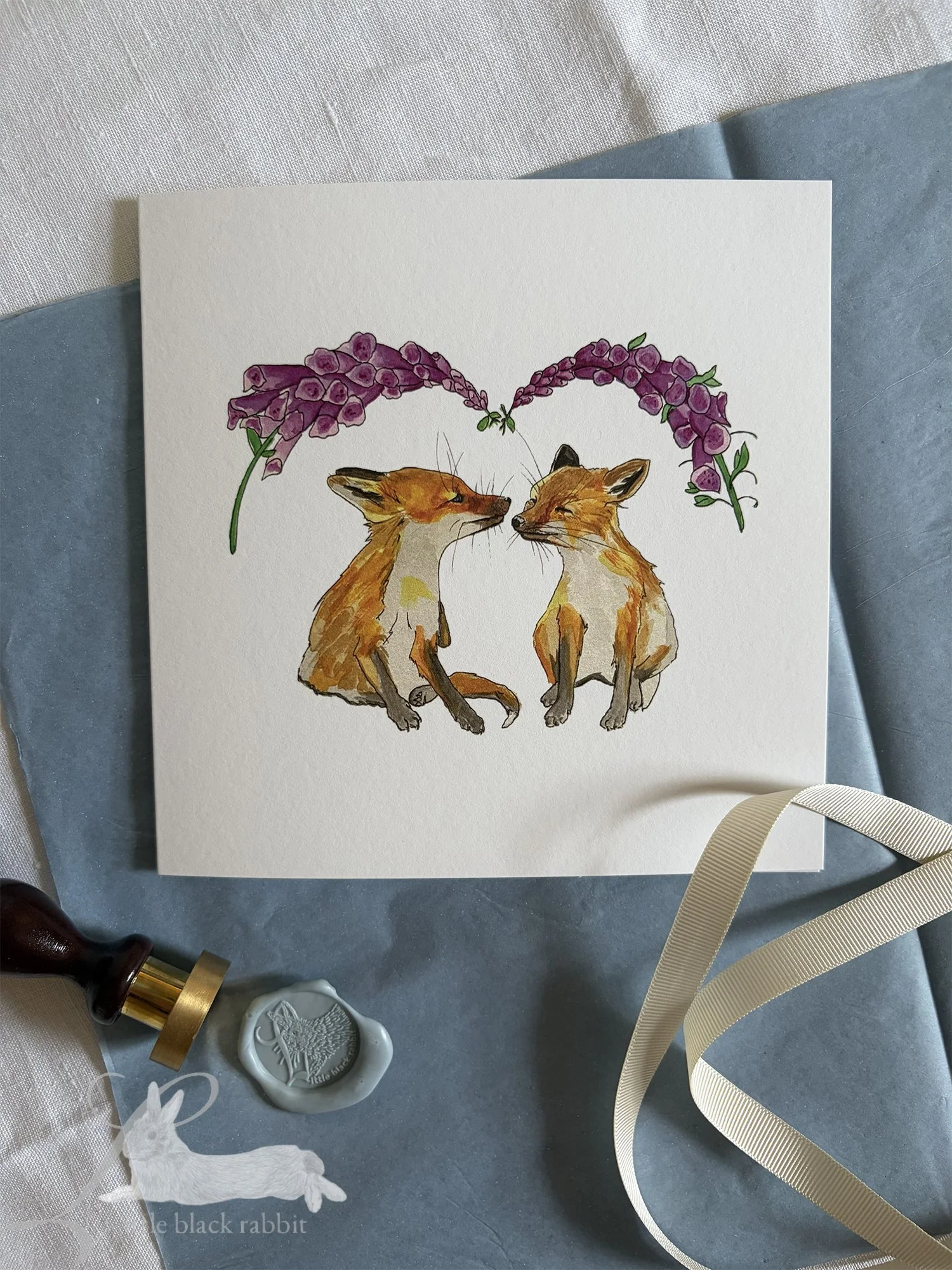 Greetings Card with two foxcubs under the foxgloves sitting on a table with a white linen table cloth and some blue tissue paper, cream ribbon and a blue wax seal with the Little Black Rabbit logo.