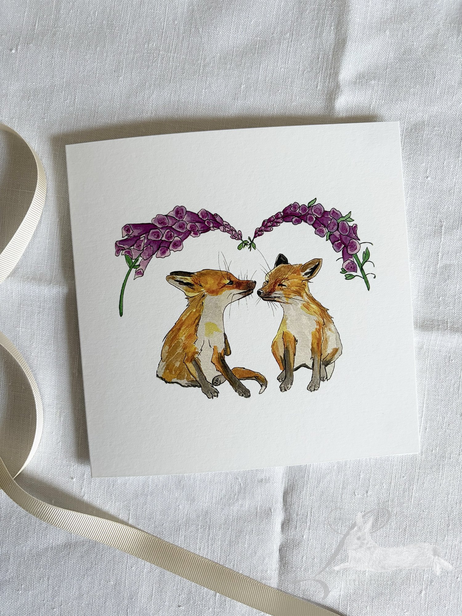 Greetings Card with two foxcubs under the foxgloves sitting on a table with a white linen table cloth and some cream ribbon..