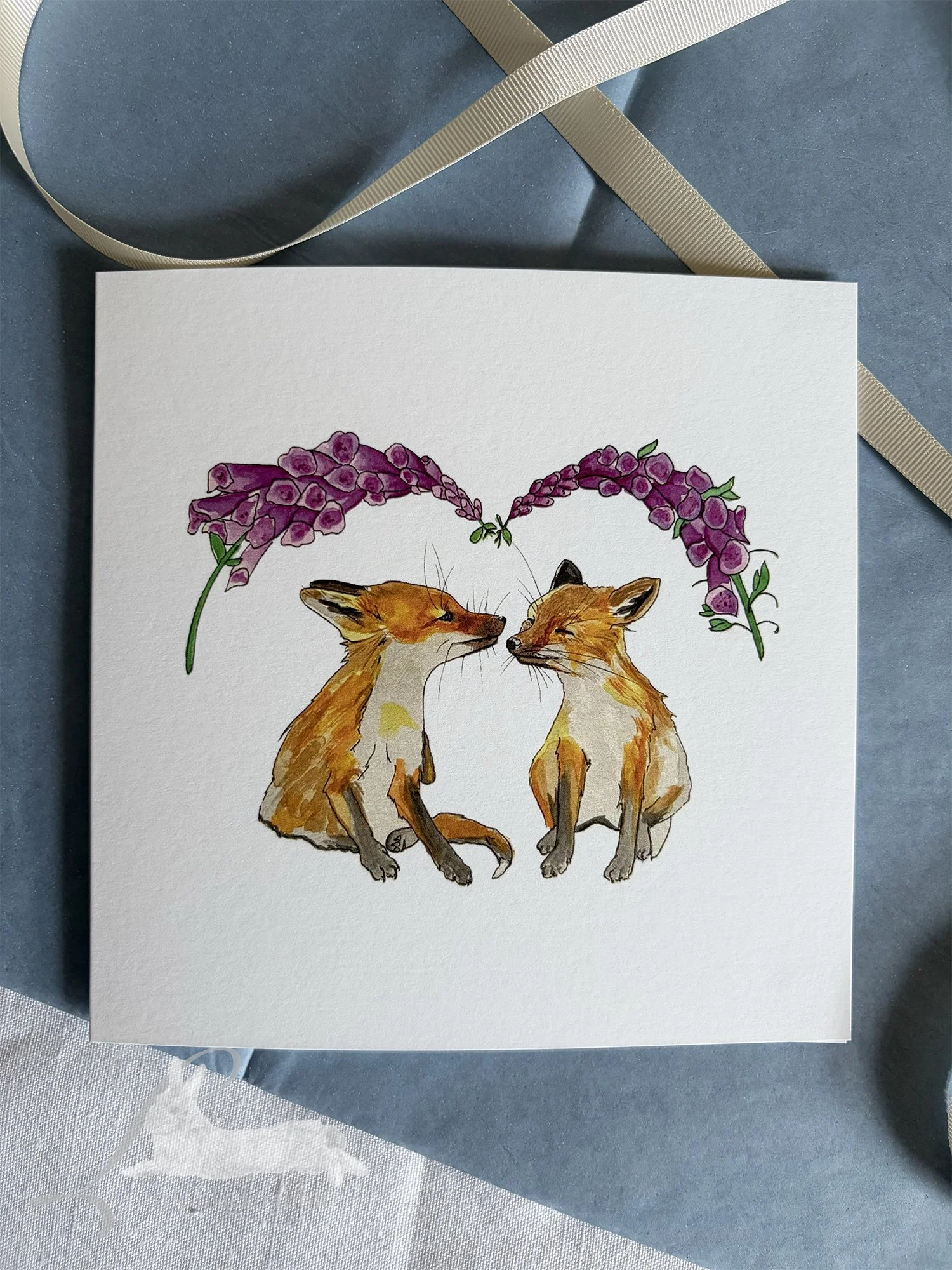 Greetings Card with two foxcubs under the foxgloves sitting on a table with a white linen table cloth and some blue tissue paper and cream ribbon.