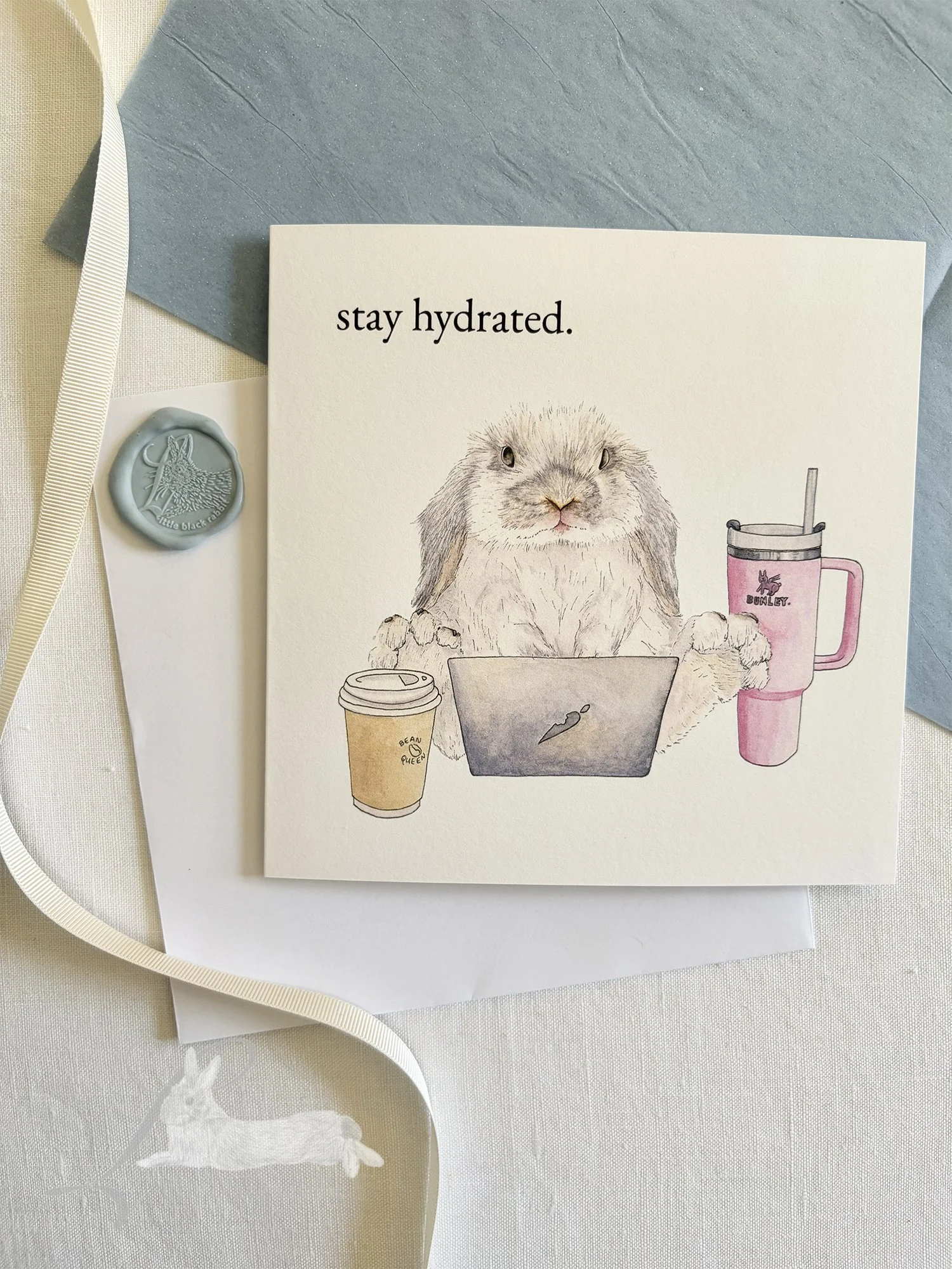 Greeting card showing a hand painted rabbit with a laptop, takeaway coffee, and waterbottle, sitting on a white linen tablecloth with cream ribbon and wax seal.