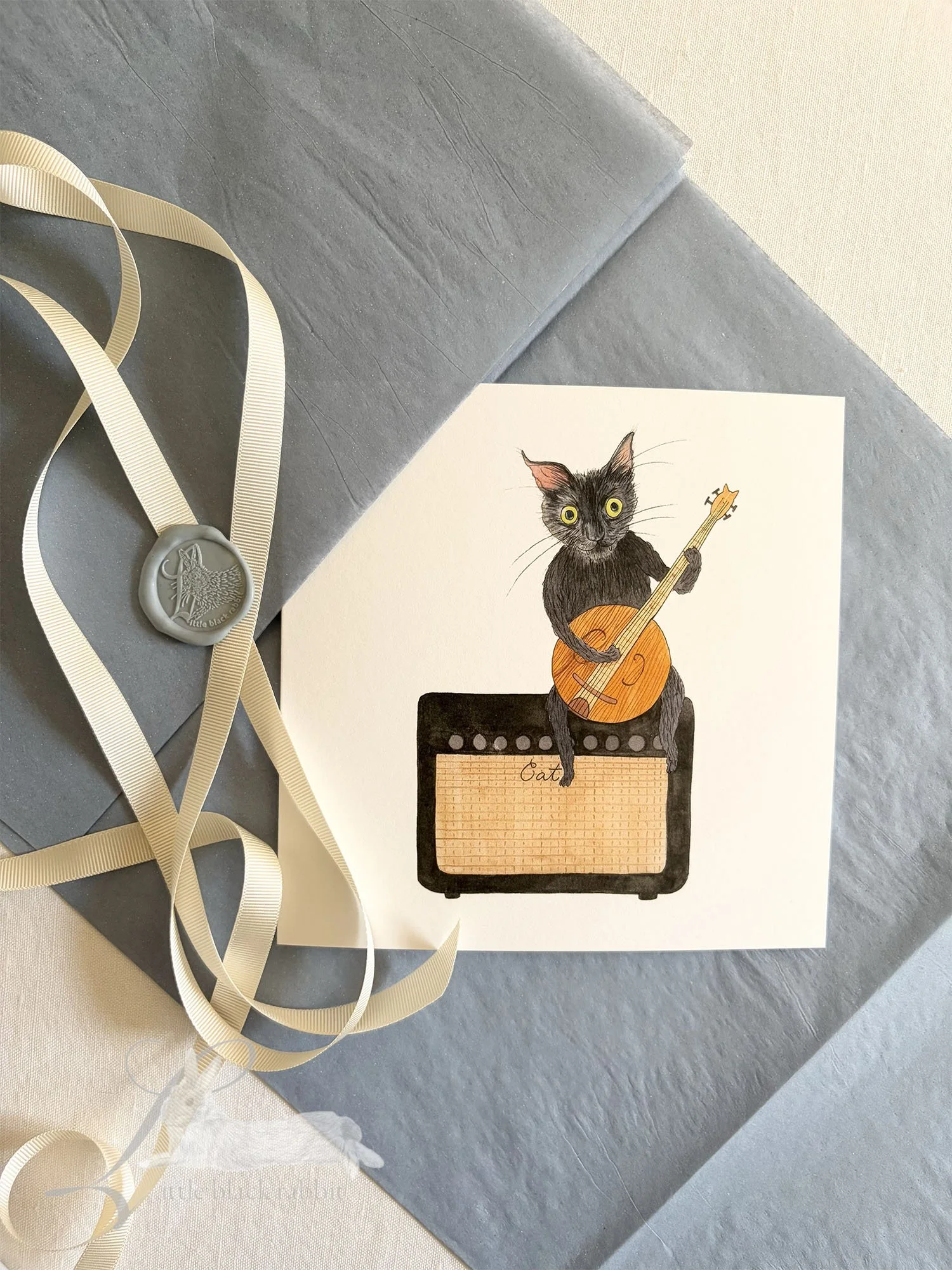 Hand painted greetings card showing a black cat with green eyes sitting on an amplifyer playing a banjo, sitting on a white linen tablecloth with blue tissue paper, cream ribbon and a wax seal.