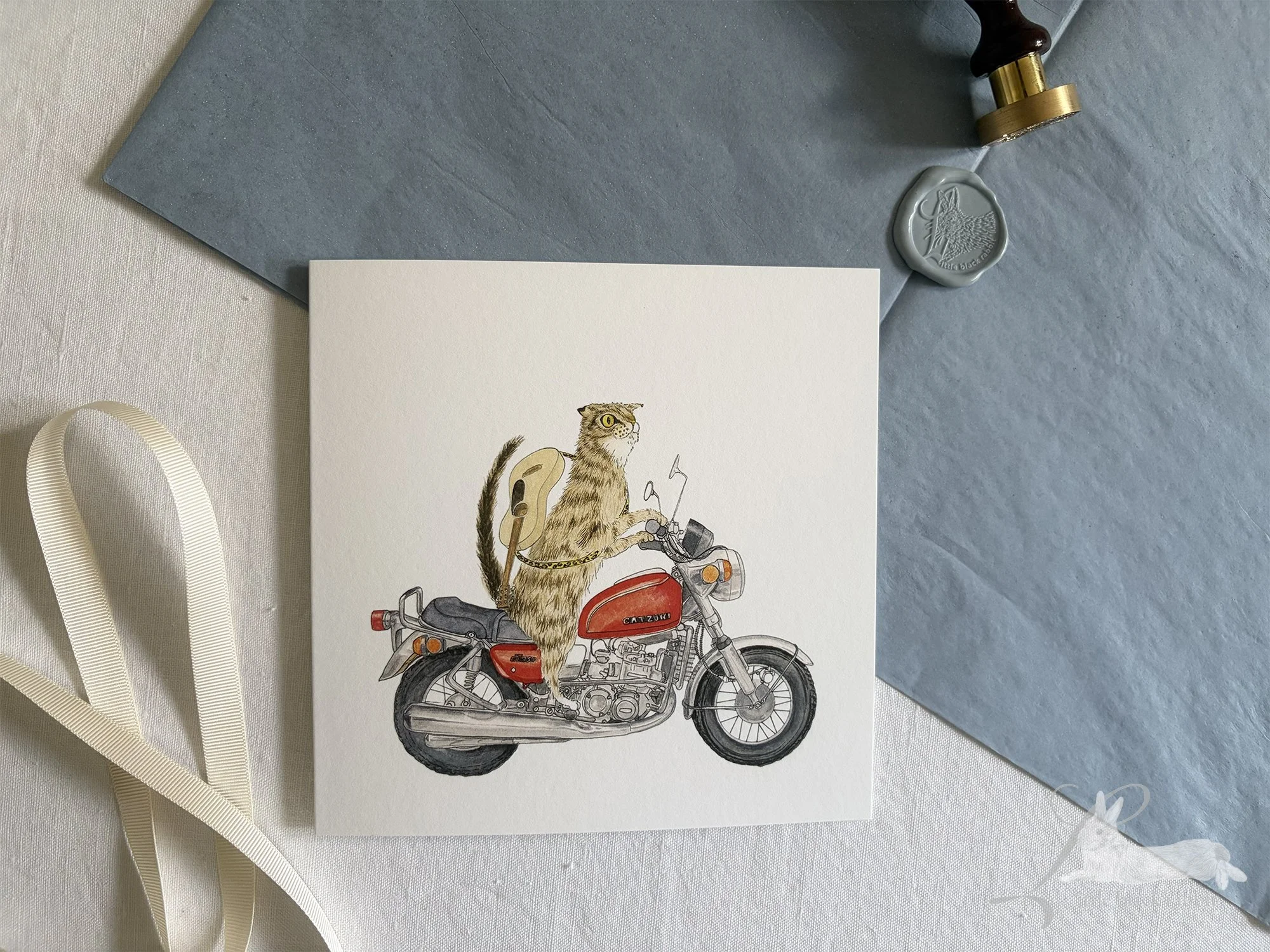 Hand painted greetings card showing a tabby cat on a red motorbike with a guitar on his back, sitting on a white linen tablecloth with cream ribbon, blue tissue paper, and a wax seal with stamp.