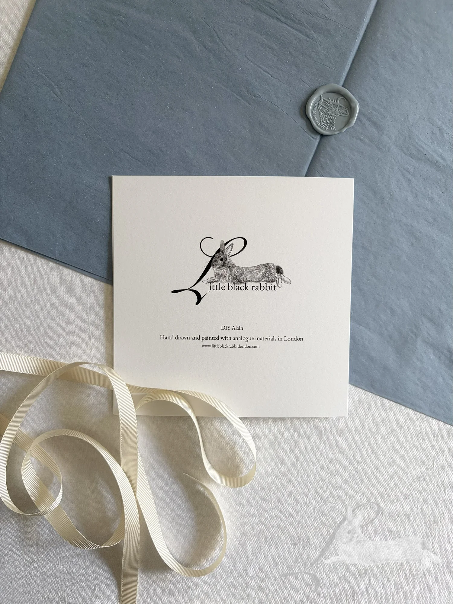 Reverse side of a hand painted greeting card showing the Little Black Rabbit logo, sitting on a white linen tablecloth with blue tissue paper, cream ribbon and a wax seal.