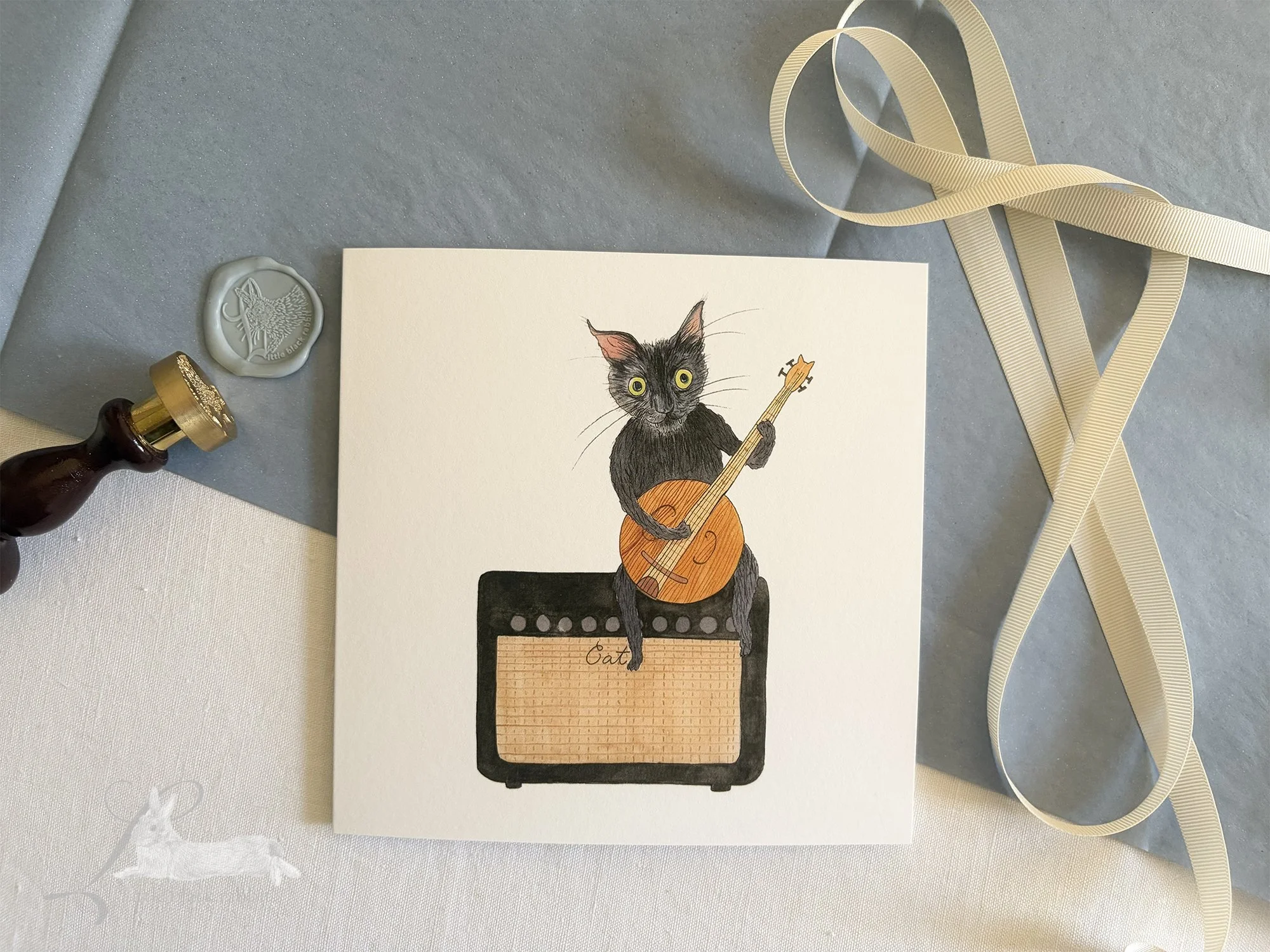Hand painted greetings card showing a black cat with green eyes sitting on an amplifyer playing a banjo, sitting on a white linen tablecloth with blue tissue paper, cream ribbon and a wax seal.