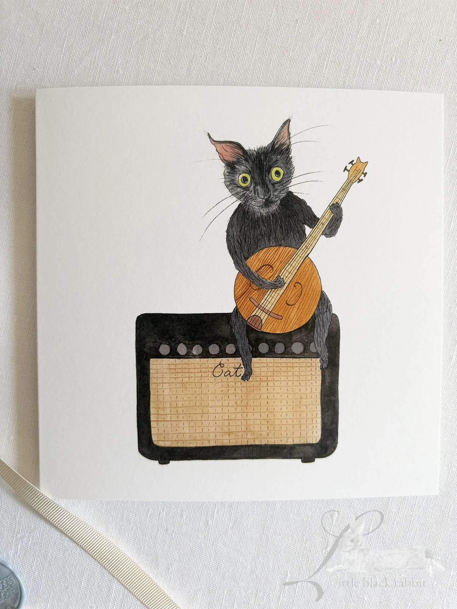 Hand painted greetings card showing a black cat with green eyes sitting on an amplifyer playing a banjo, sitting on a white linen tablecloth.