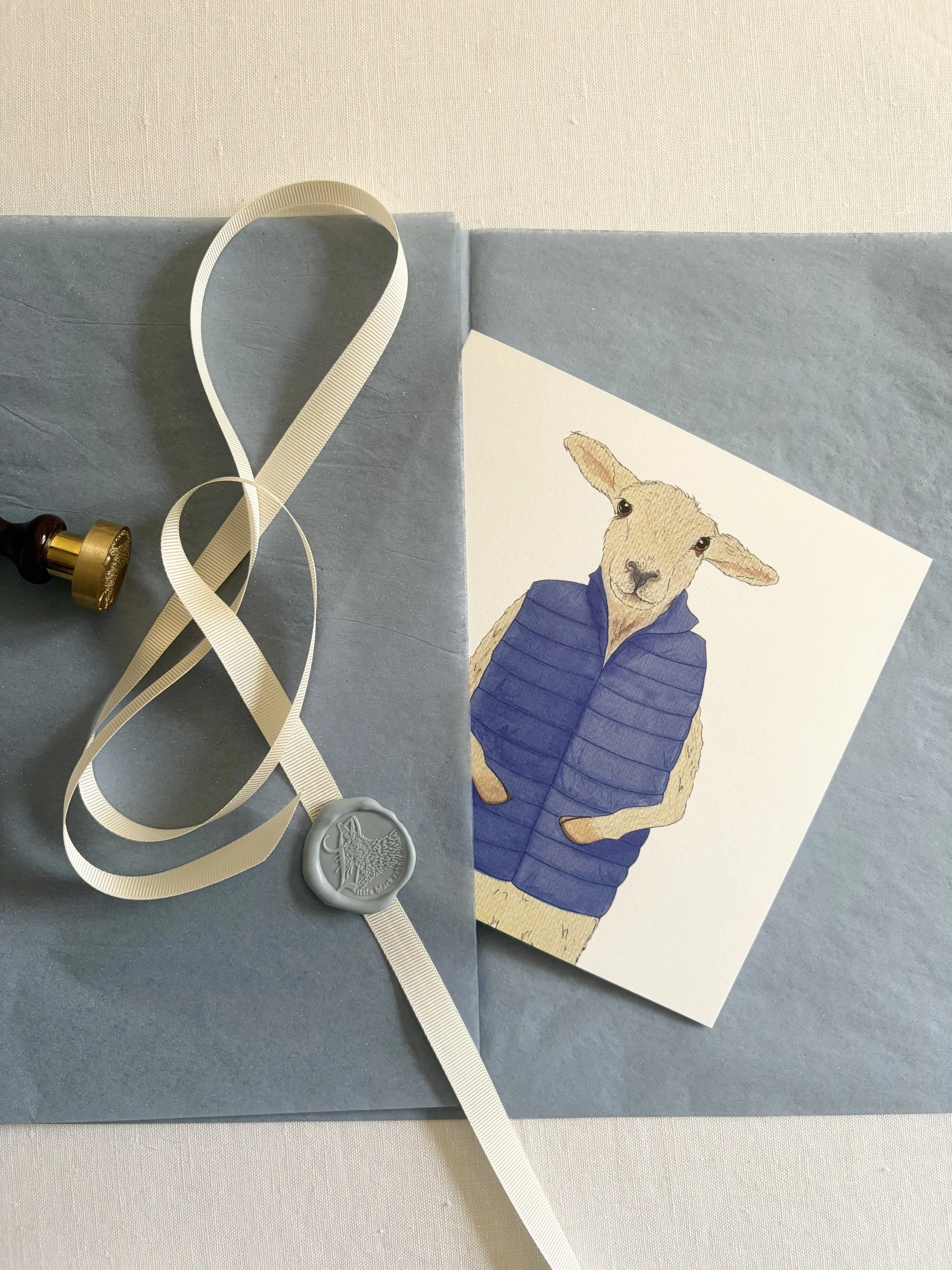 Handpainted greeting card featuring a smiling sheep in a blue quilted gilet, sitting on a white linen tablecloth with blue tissue paper, cream ribbon, and a blue wax seal.