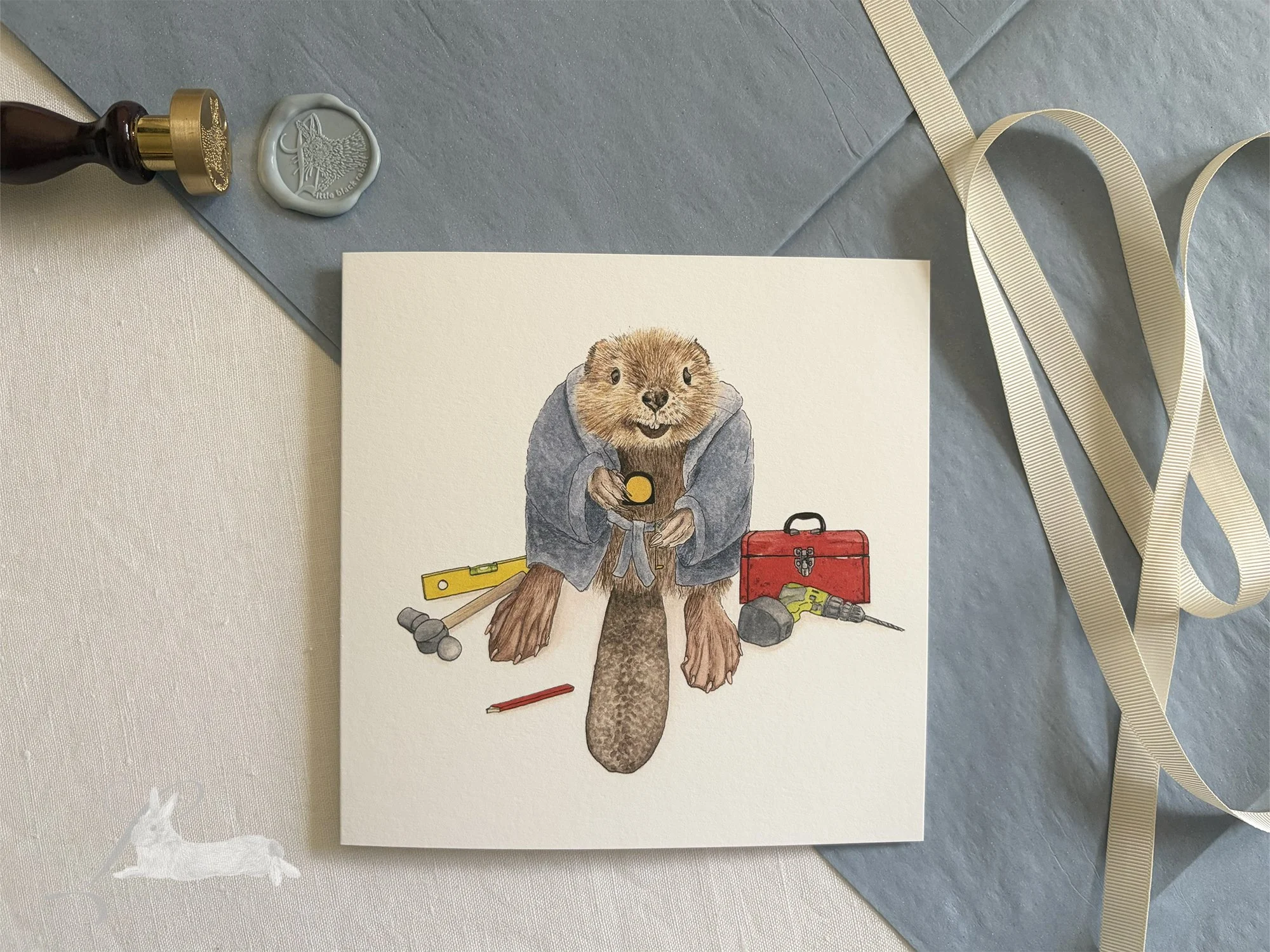 Hand painted greetings card showing a beaver in a blue dressing gown surrounded by DIY tools and a tool box, sitting on a white linen tablecloth with blue tissue paper, cream ribbon and a wax seal.