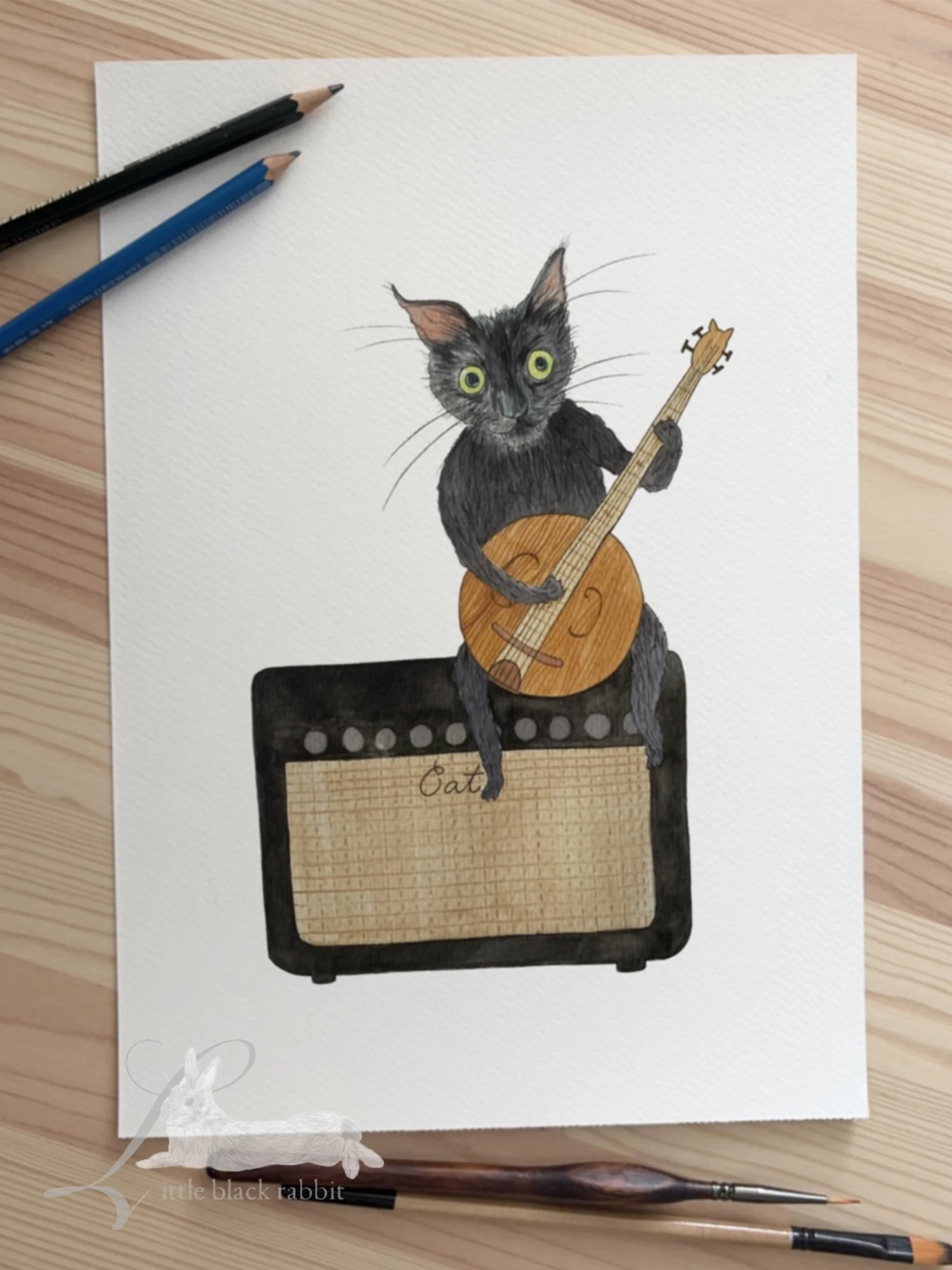 Hand painted watercolour illustration of a black cat with green eyes sitting on an amplifyer swinging his legs, sitting on a wooden table with a tube of paint and a paintbrush.