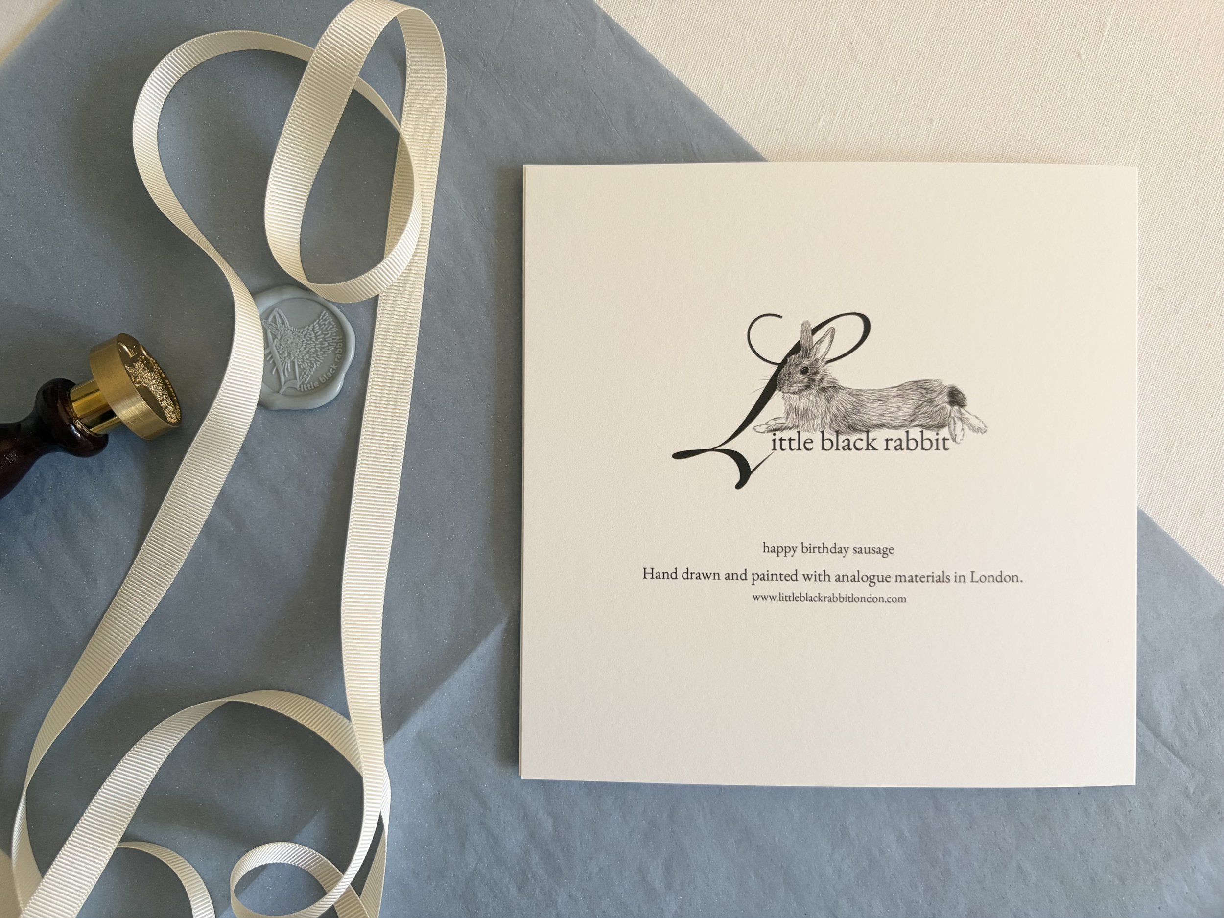 Zoomed out image of a birthday card sitting face down showing the Little Black Rabbit logo on a table with dusty blue tissue paper, a white linen tablecloth, and cream ribbon.